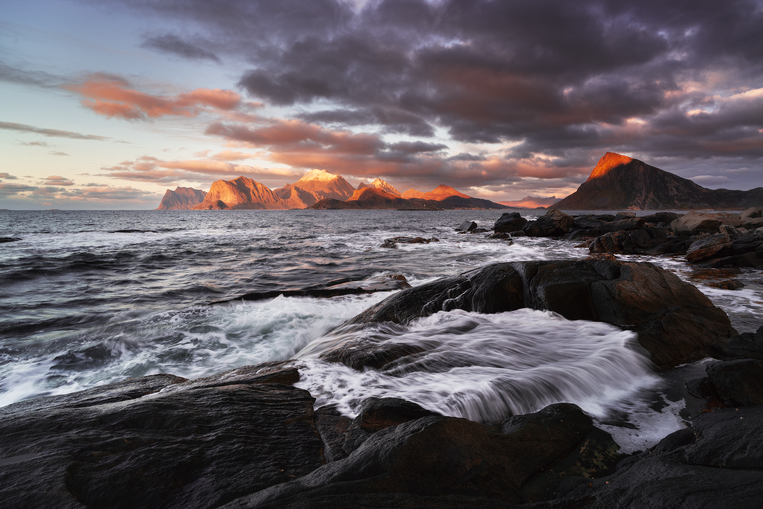 Scenic view of rocky coastline with waves crashing, mountains in the background, and dramatic clouds in the sky during sunset.