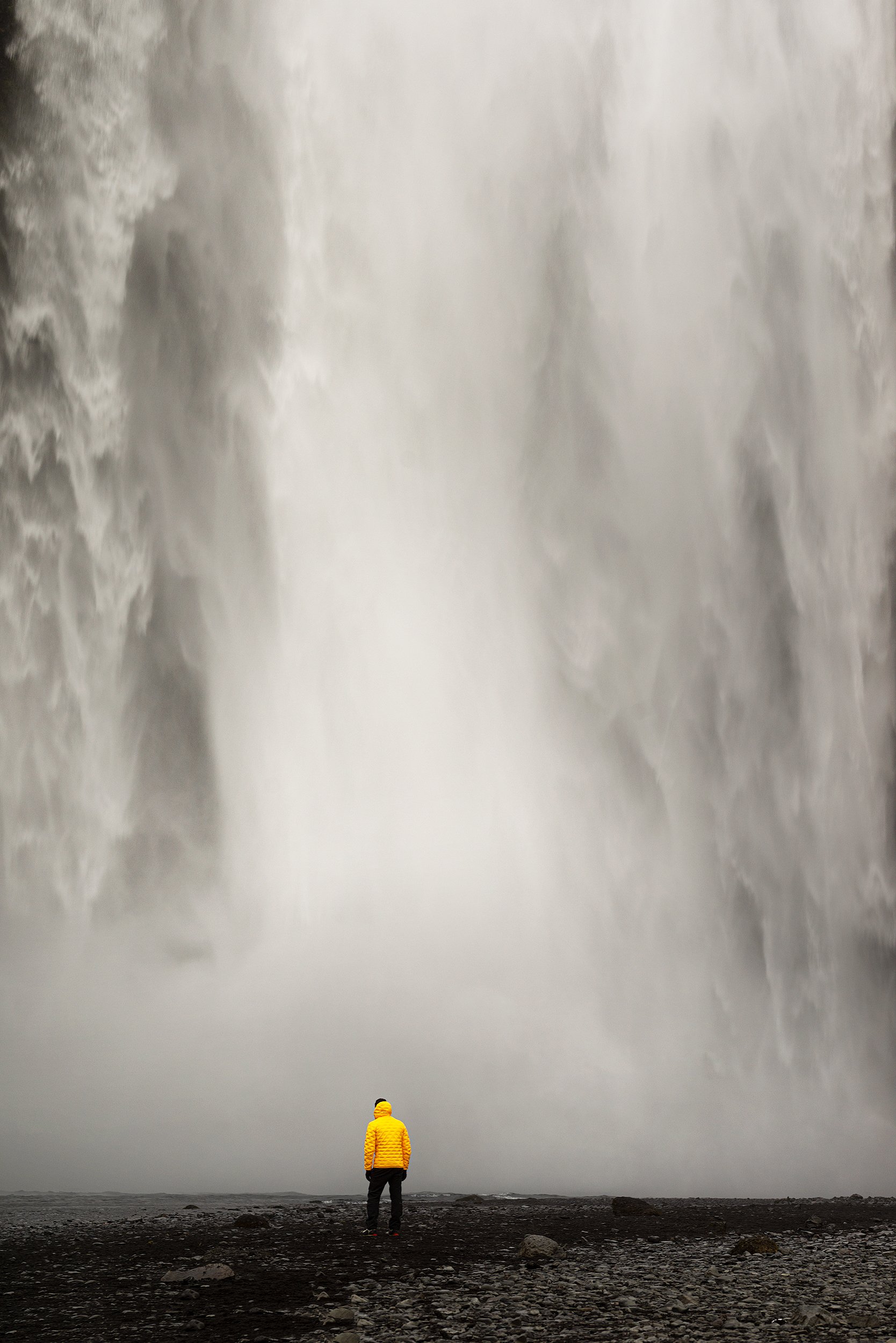 Person in a yellow jacket and dark pants standing on rocky ground, facing a massive waterfall.