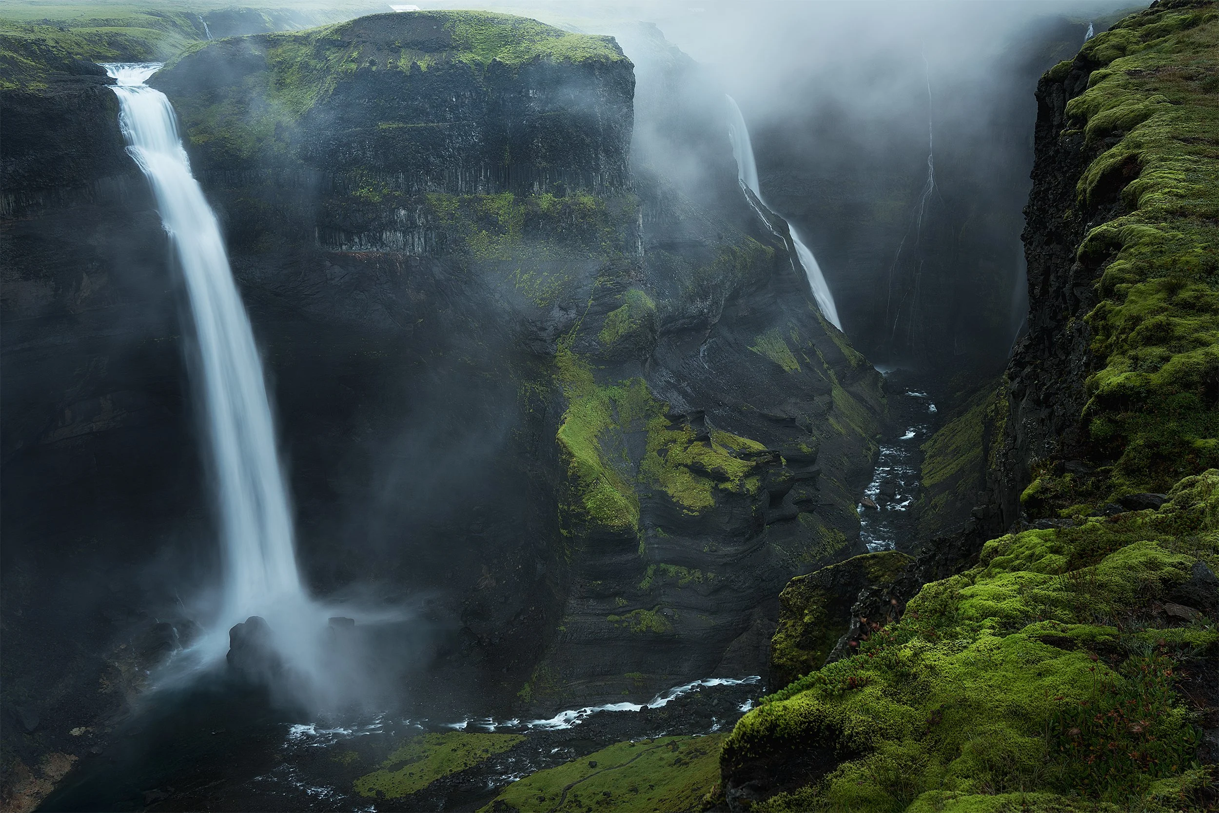 Misty waterfall cascading down black volcanic rocks surrounded by lush green moss and vegetation in a deep canyon.