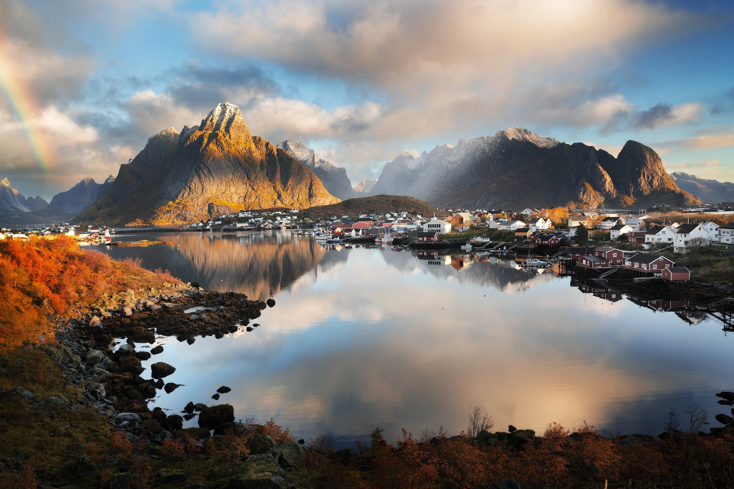 Scenic landscape of a coastal village with colorful houses, surrounded by tall mountain peaks, a calm reflective body of water, and a rainbow in the sky
