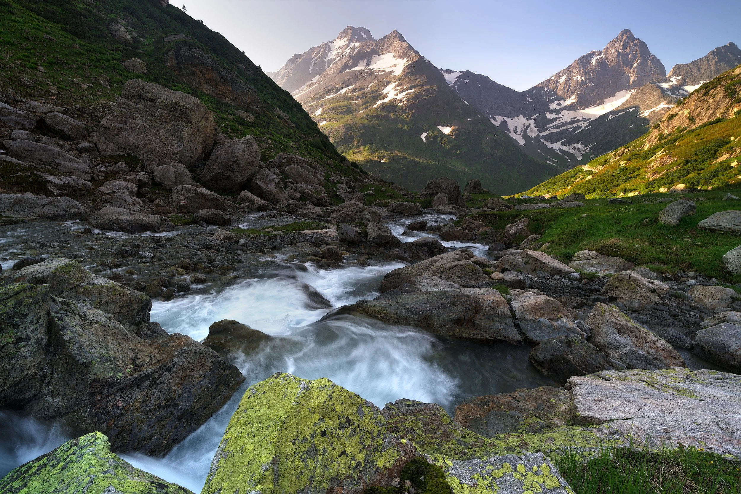 A mountain stream flowing through a rocky valley with snow-capped peaks in the background and green vegetation along the banks.