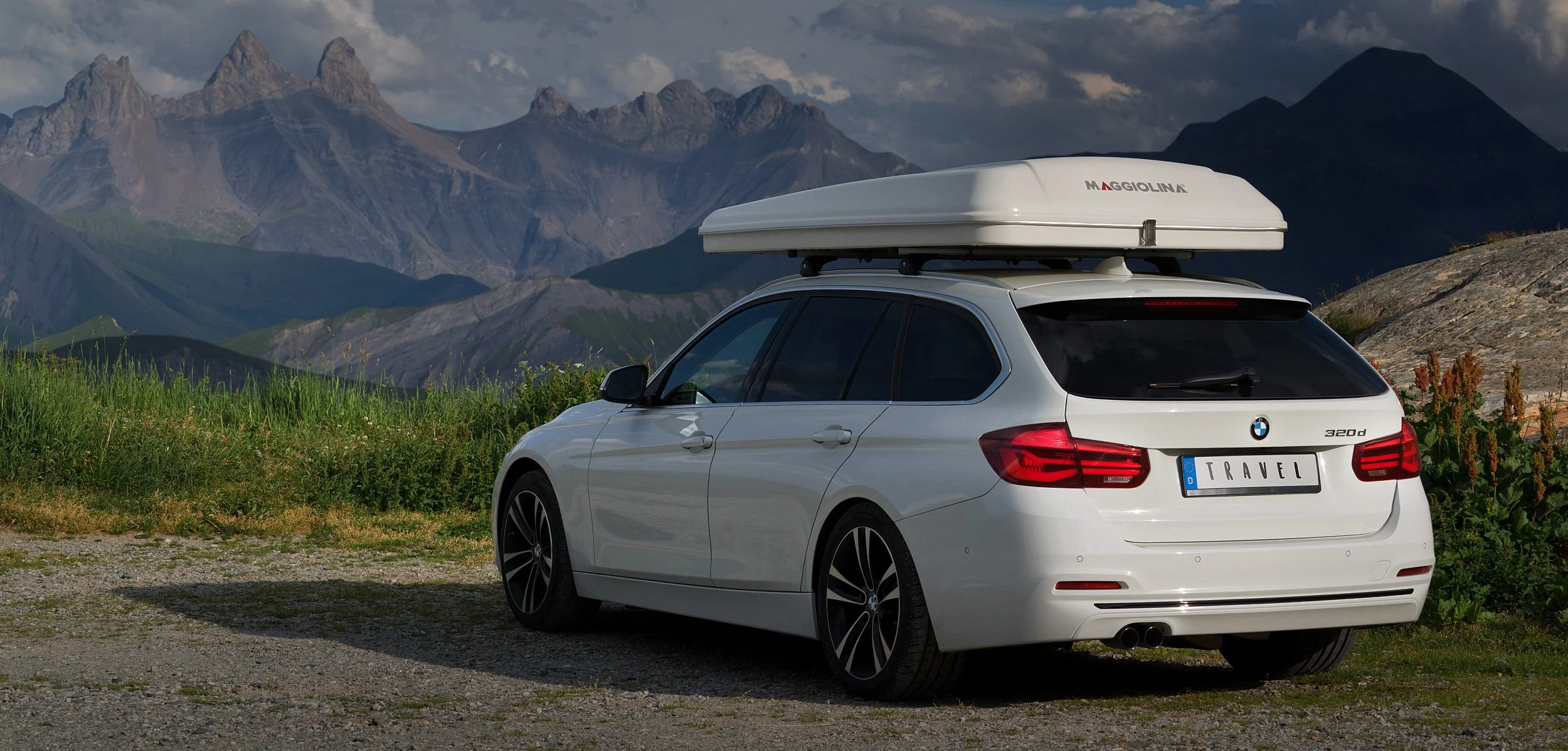White BMW station wagon with a rooftop cargo box parked on a gravel area near green grass and mountains in the background.