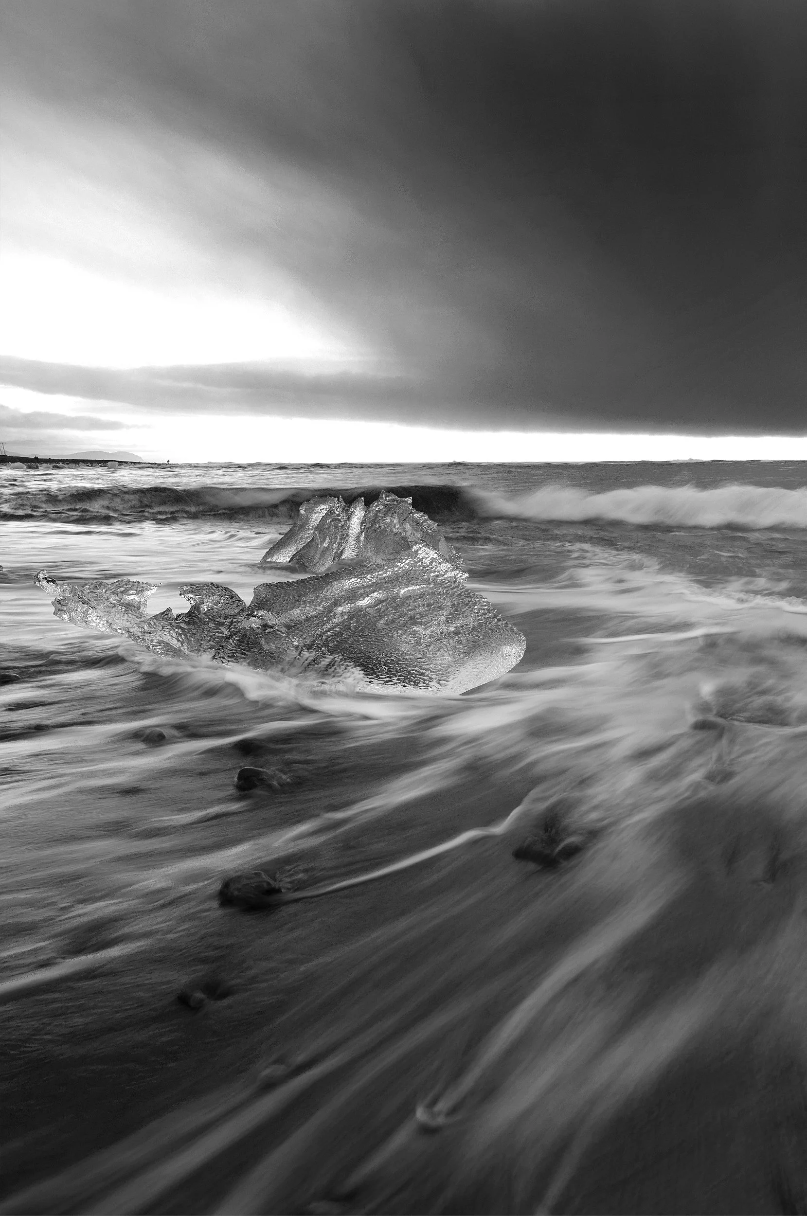 Black and white photograph of the ocean with waves crashing on rocks in the foreground, and dark clouds overhead.