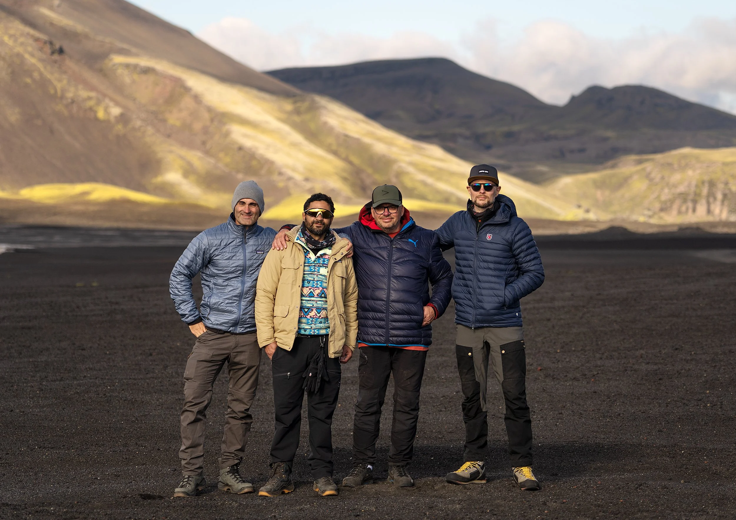 Four men in outdoor jackets and hats standing on volcanic black sand with mountains in the background.