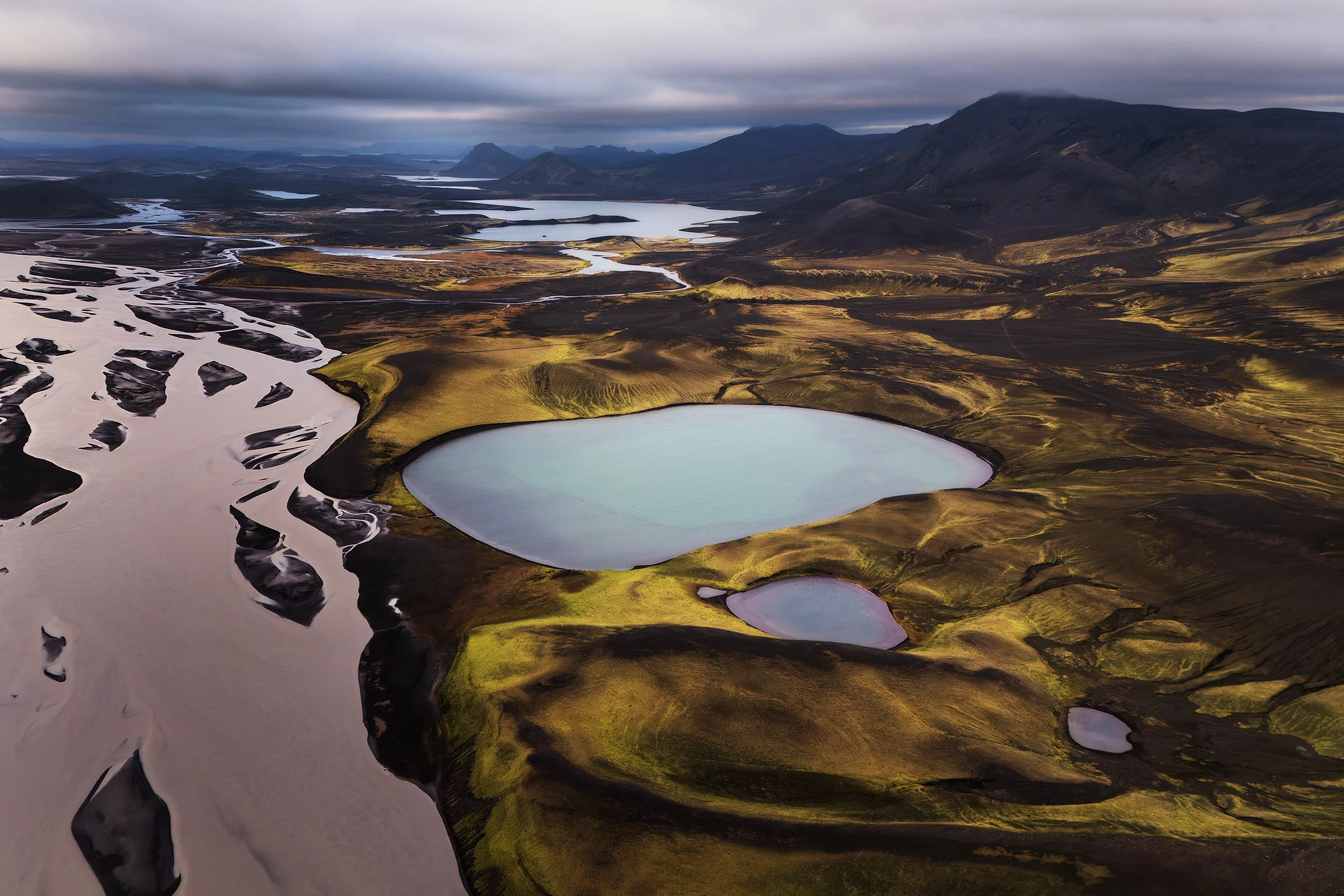 Aerial view of a volcanic landscape with multiple crater lakes, black sand, and moss-covered terrain under a cloudy sky in Iceland.