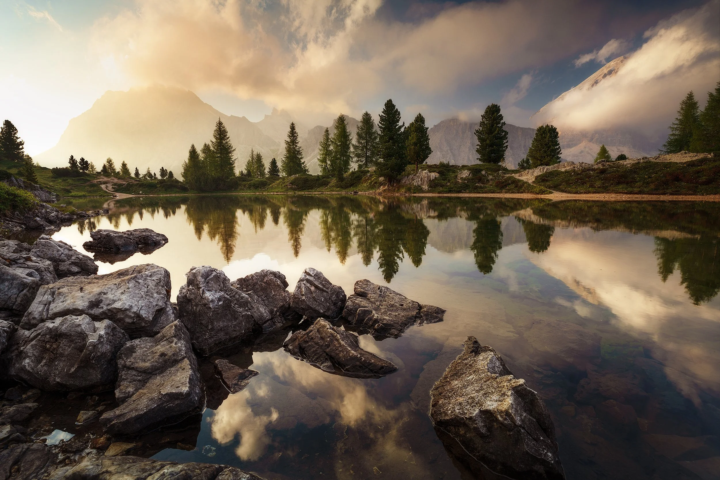 Scenic mountain landscape with a calm lake, rocks in the foreground, evergreen trees, mountains in the background, and clouds in the sky during sunset.