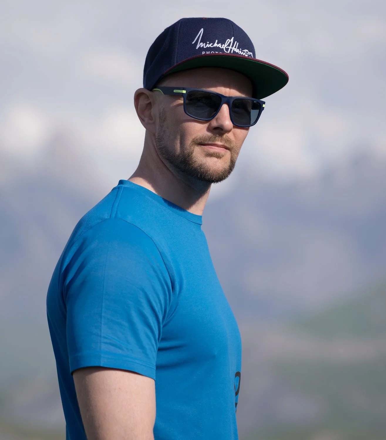 A man wearing sunglasses, a blue t-shirt, and a dark baseball cap with white writing, standing outdoors with a cloudy sky in the background.