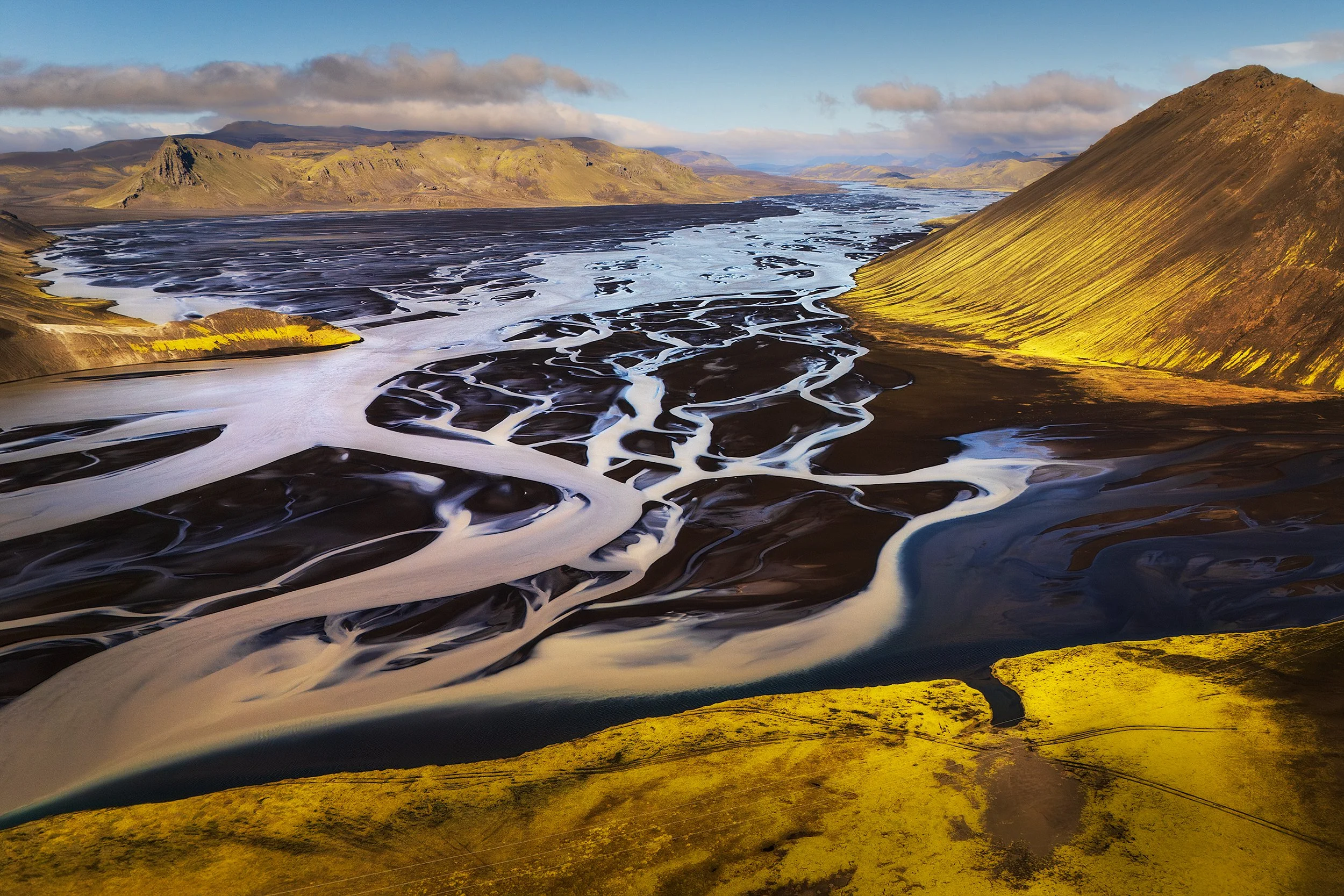 Aerial view of a black sand river with white braided channels flowing through a mountainous landscape with green and brown slopes, under a partly cloudy sky.