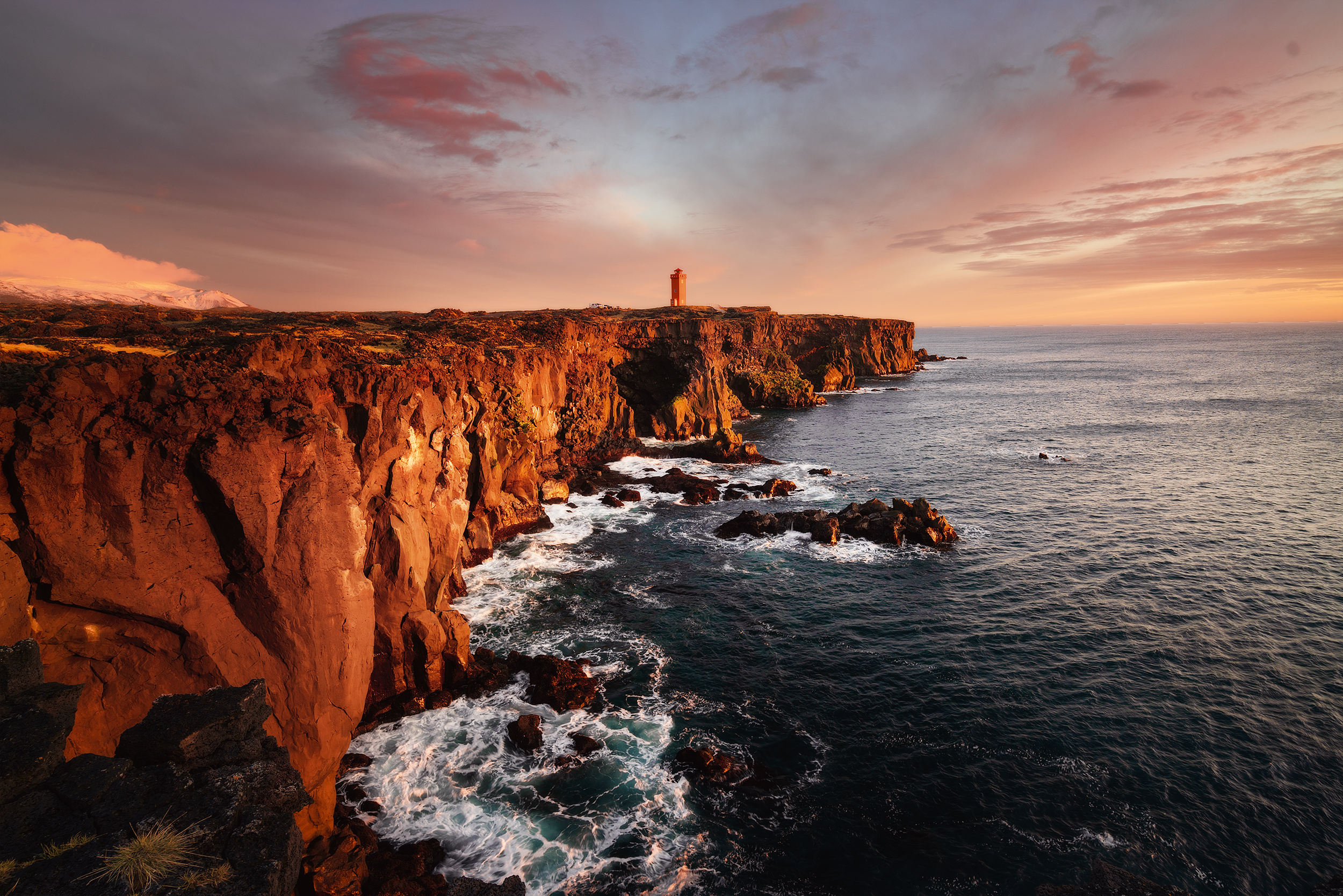 Sunset over rugged coastal cliffs with a lighthouse on top, waves crashing against rocks below, and a colorful sky with scattered clouds.