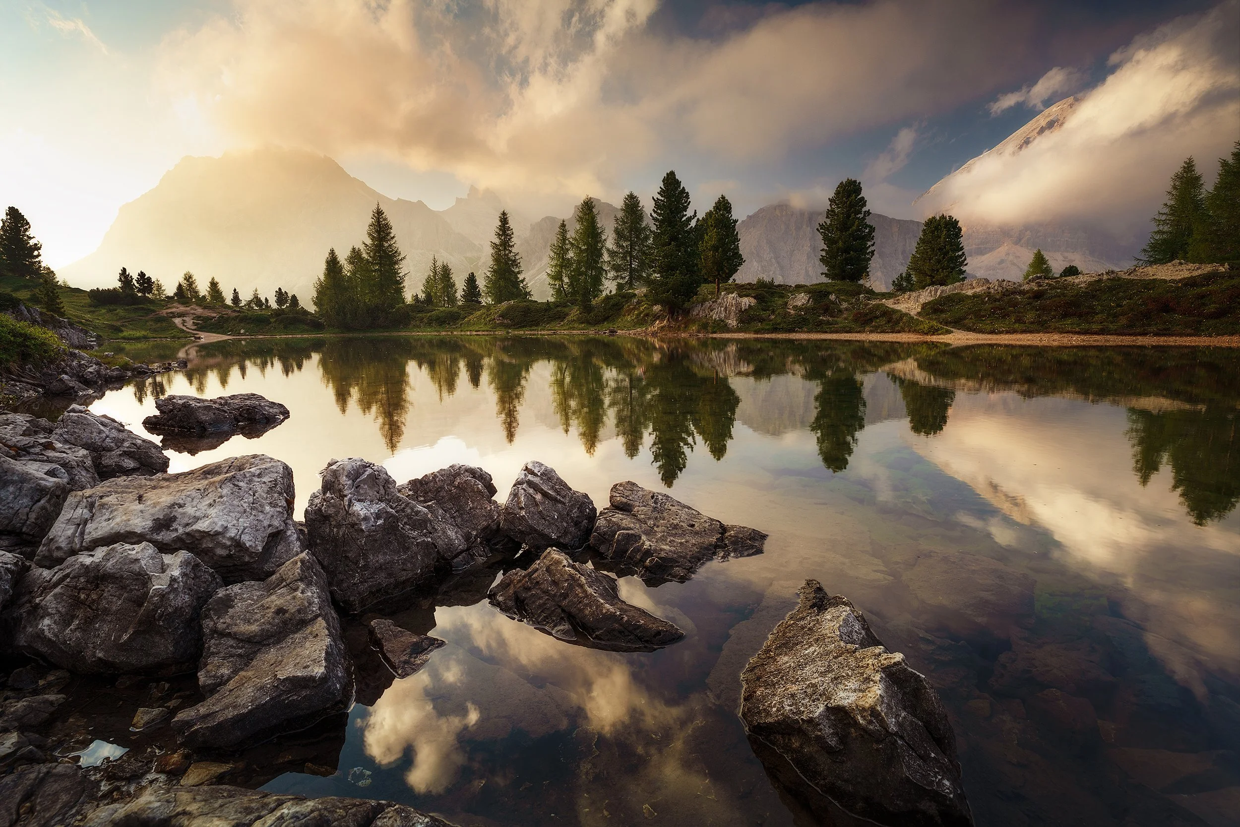 A serene mountain lake scene with rocky shore in the foreground, calm water reflecting pine trees, cloudy sky, and distant mountains.