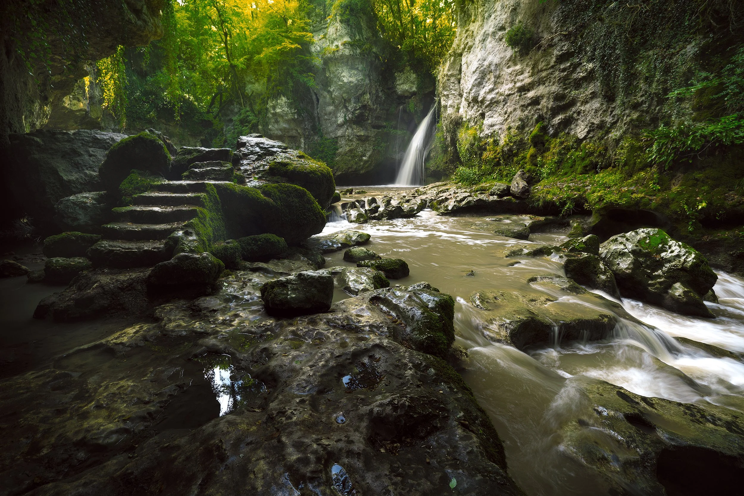 A small waterfall cascading into a rocky stream surrounded by lush green trees and moss-covered rocks in a forest