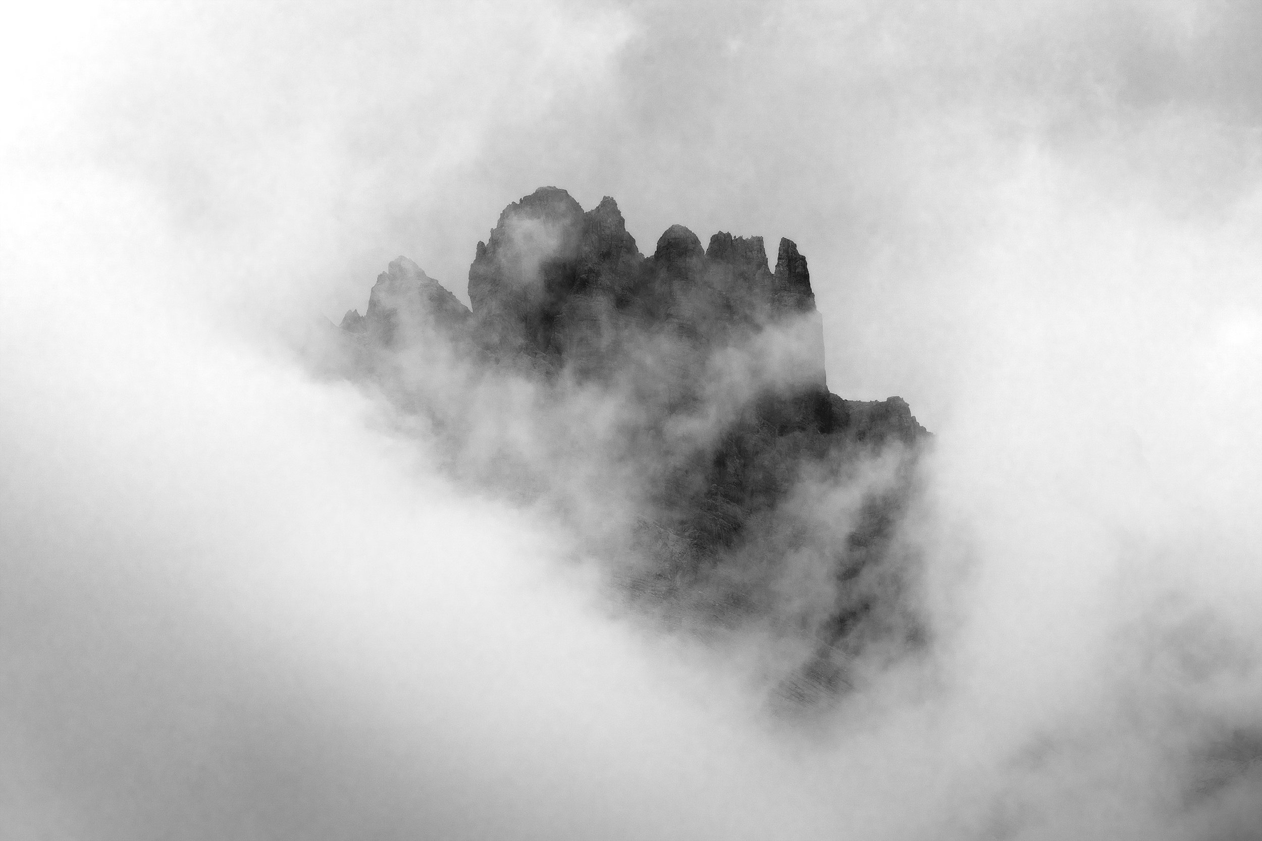 Black and white photograph of a mountain peak partially obscured by clouds, with mist or fog surrounding the base.
