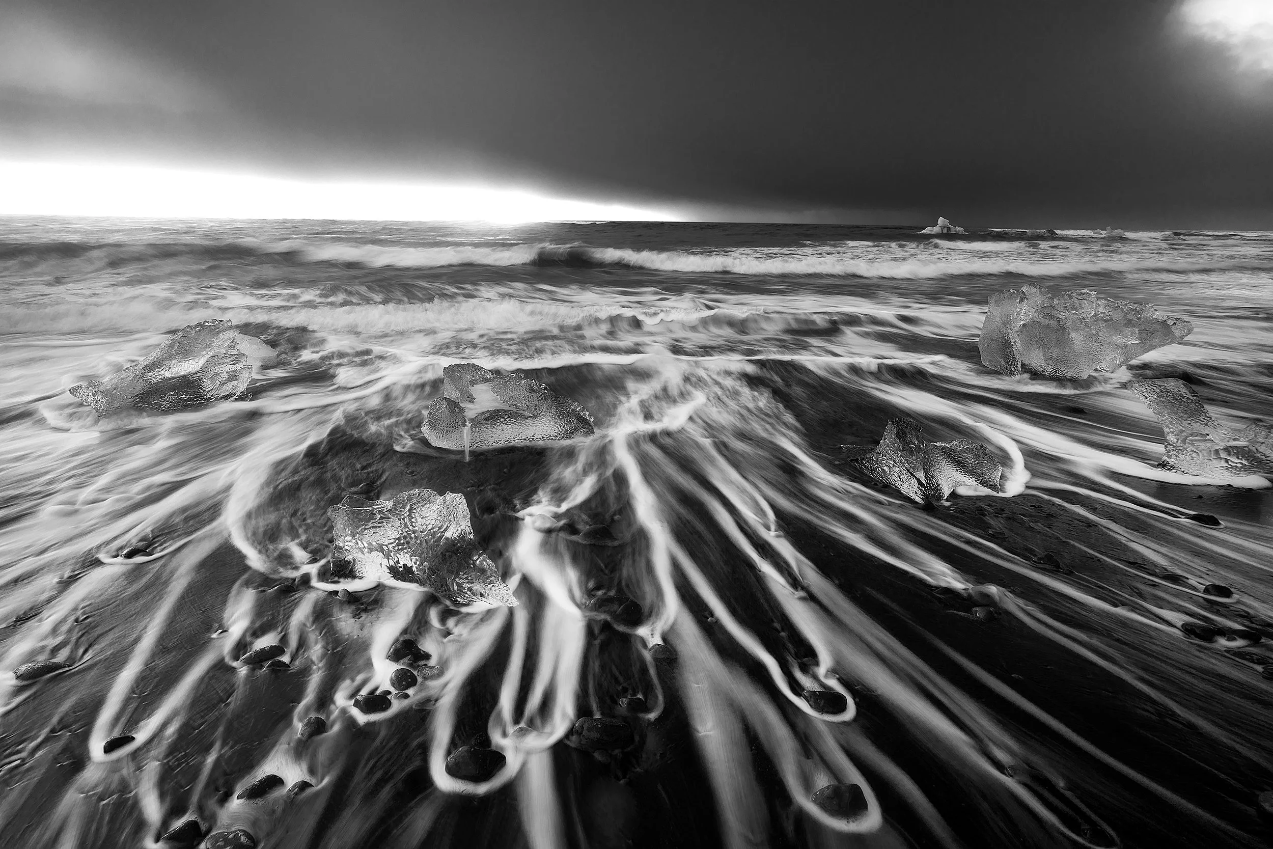 Black and white photo of waves crashing on a beach with chunks of ice scattered on the shore, dark stormy clouds overhead.