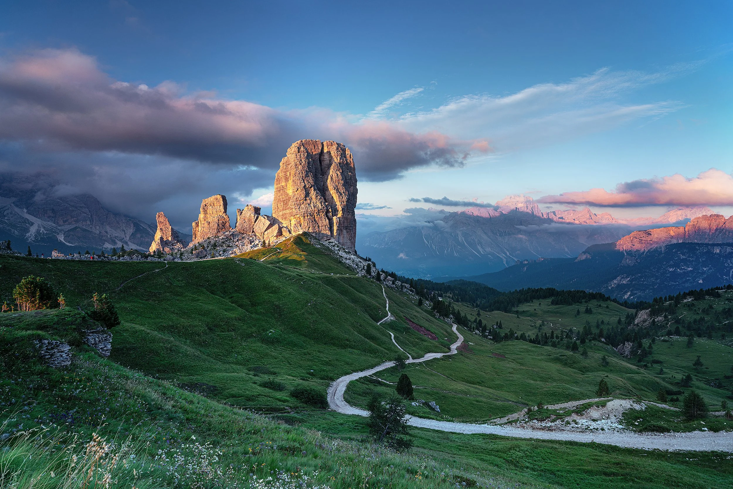 Scenic mountain landscape with green rolling hills, a winding dirt road, and tall rocky peaks under a partly cloudy sky during sunset.