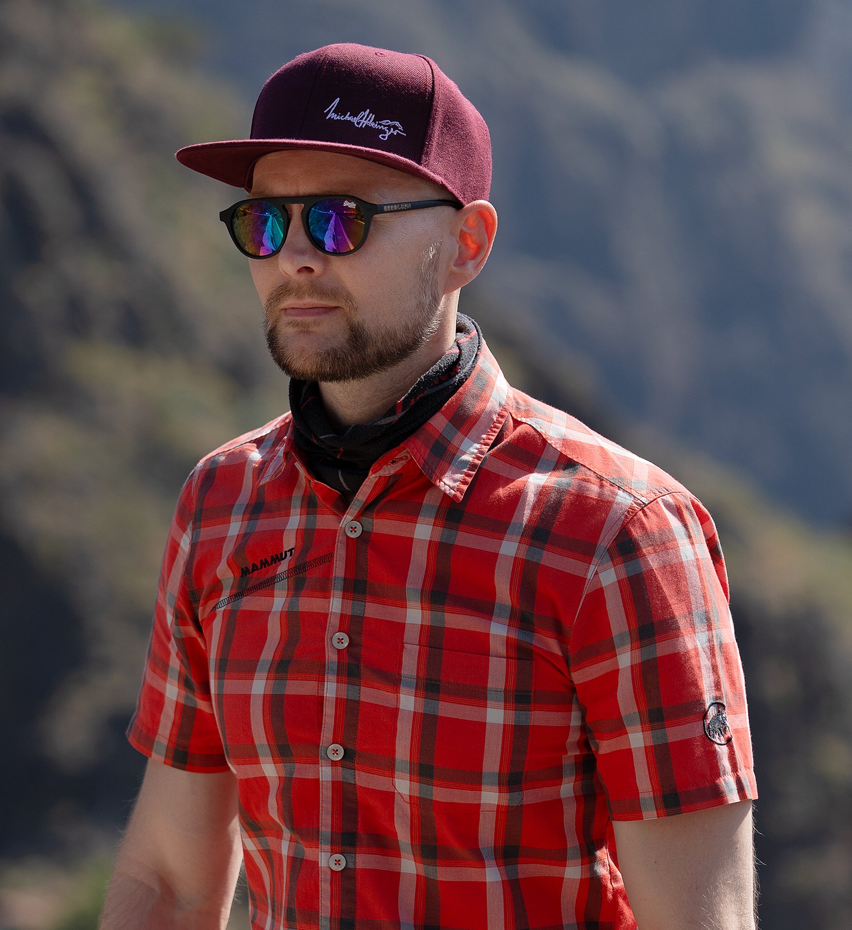 A man wearing a maroon cap, sunglasses, a red plaid shirt, and a neck gaiter outdoors with a mountainous background.
