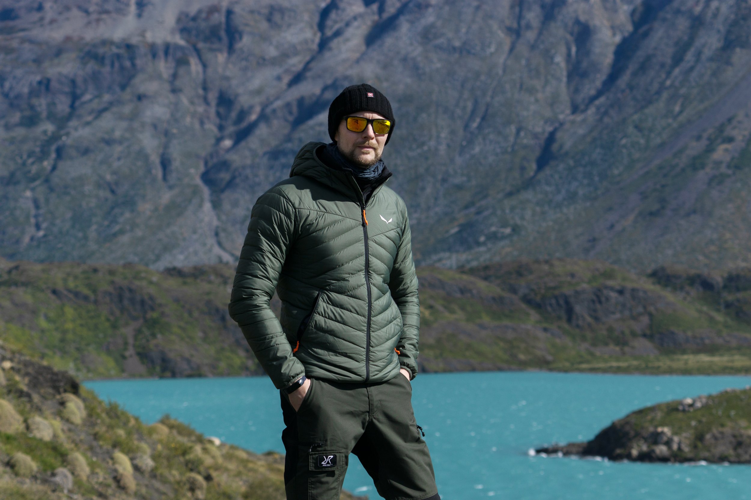 A man in a green puffy jacket, black beanie, and sunglasses standing outdoors near a lake with mountains in the background.