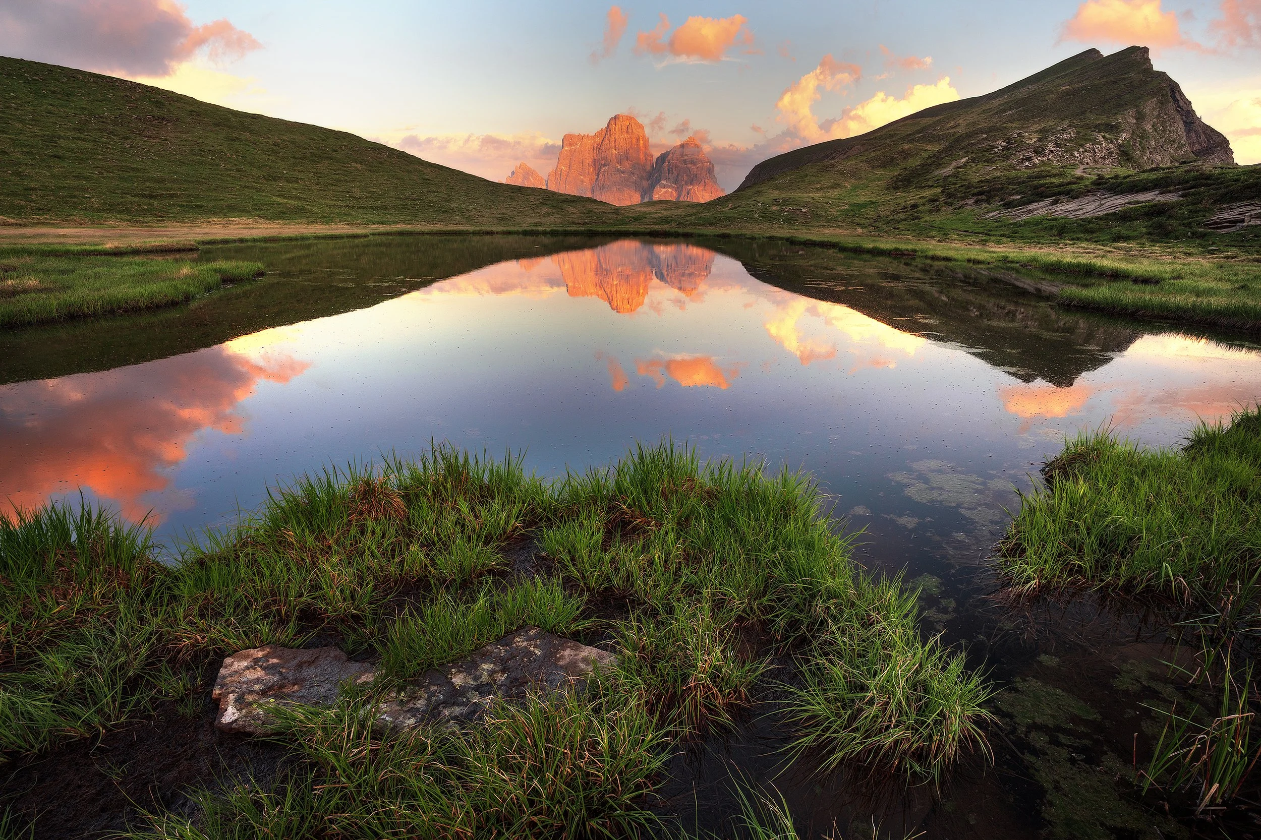 A serene landscape of a mountain valley with a calm reflective lake, green grass in the foreground, and mountains and a pastel sky with clouds in the background at sunset.