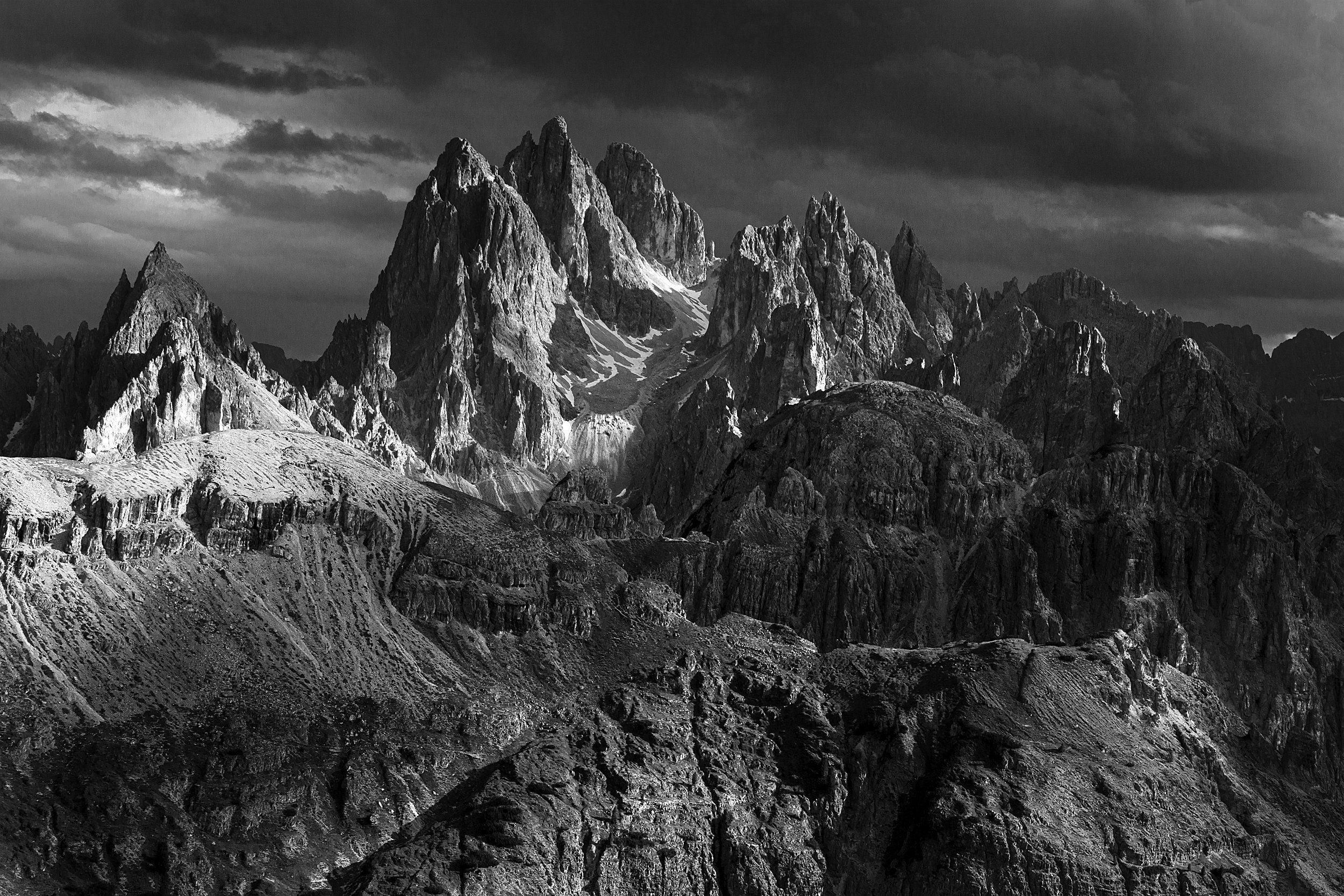 Black and white photograph of rugged mountain peaks with snow patches and dramatic cloudy sky