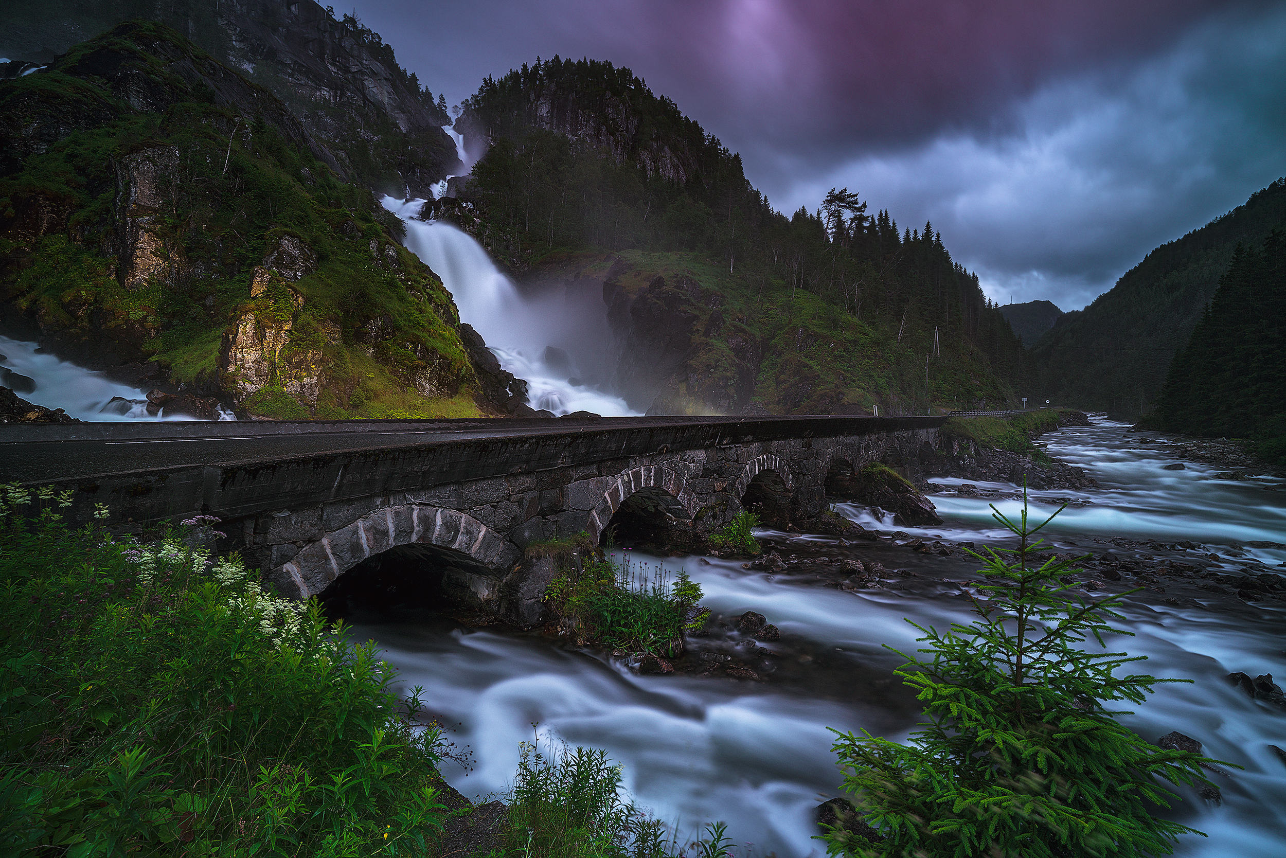 A scenic landscape of a river flowing through a valley with a stone bridge, a waterfall cascading down rocky cliffs, and a stormy sky with dark clouds surrounded by green trees and mountains.