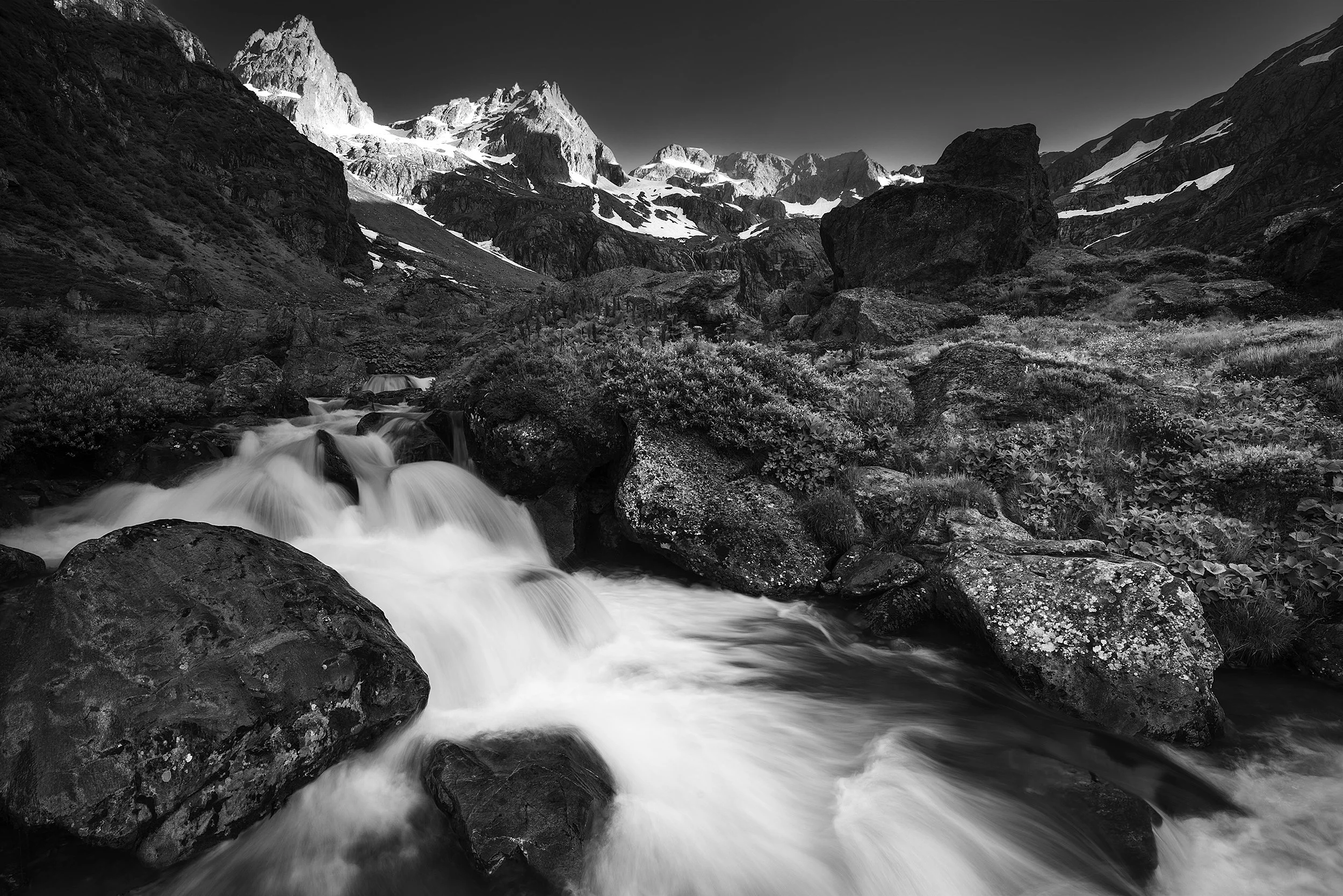 Black and white landscape of a mountain scene with a rushing stream in the foreground, rocky terrain, and snow-capped peaks in the background.