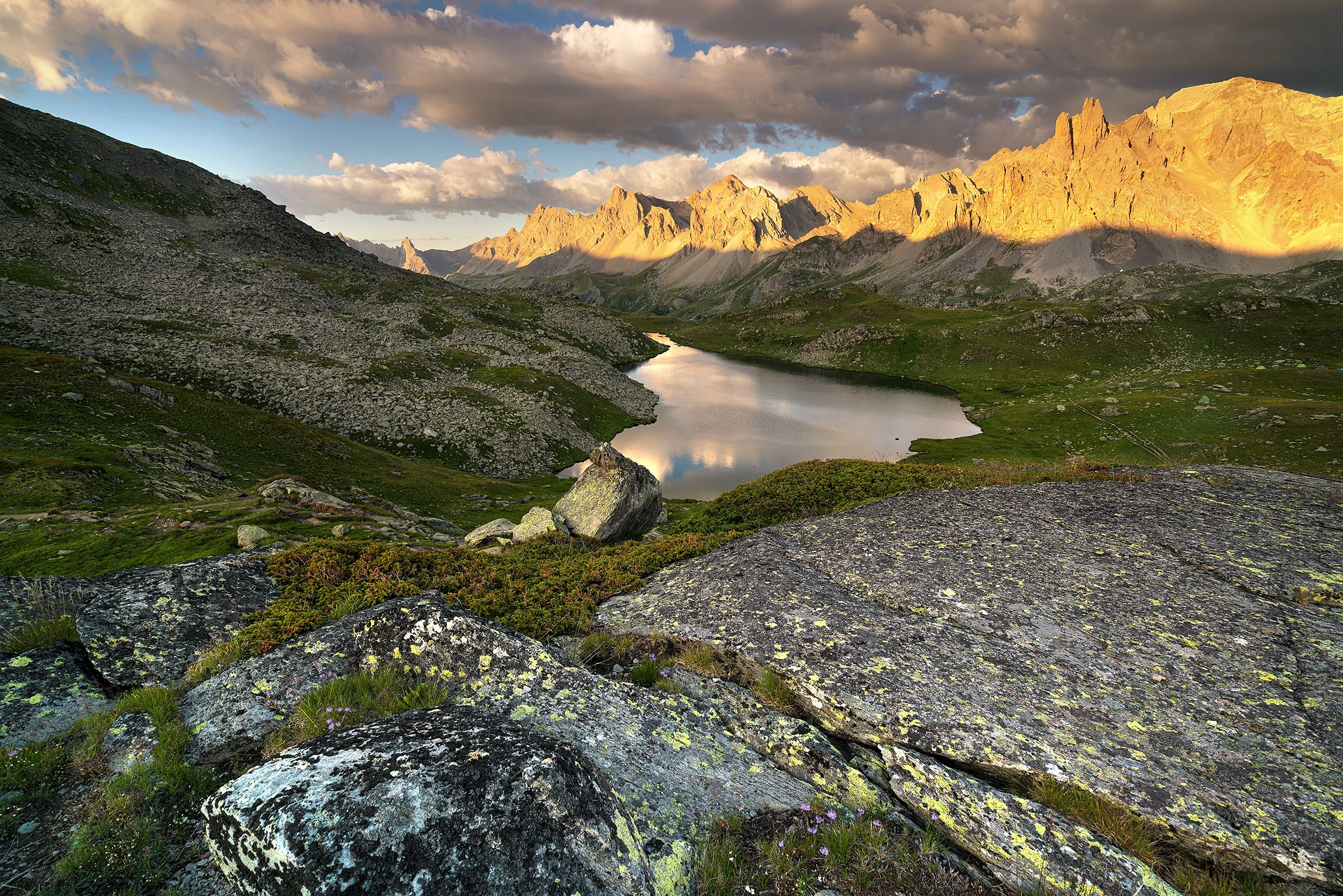 A serene mountain landscape at sunset featuring a calm lake surrounded by rocky terrain and green patches, with jagged mountains lit by golden sunlight under a partly cloudy sky.