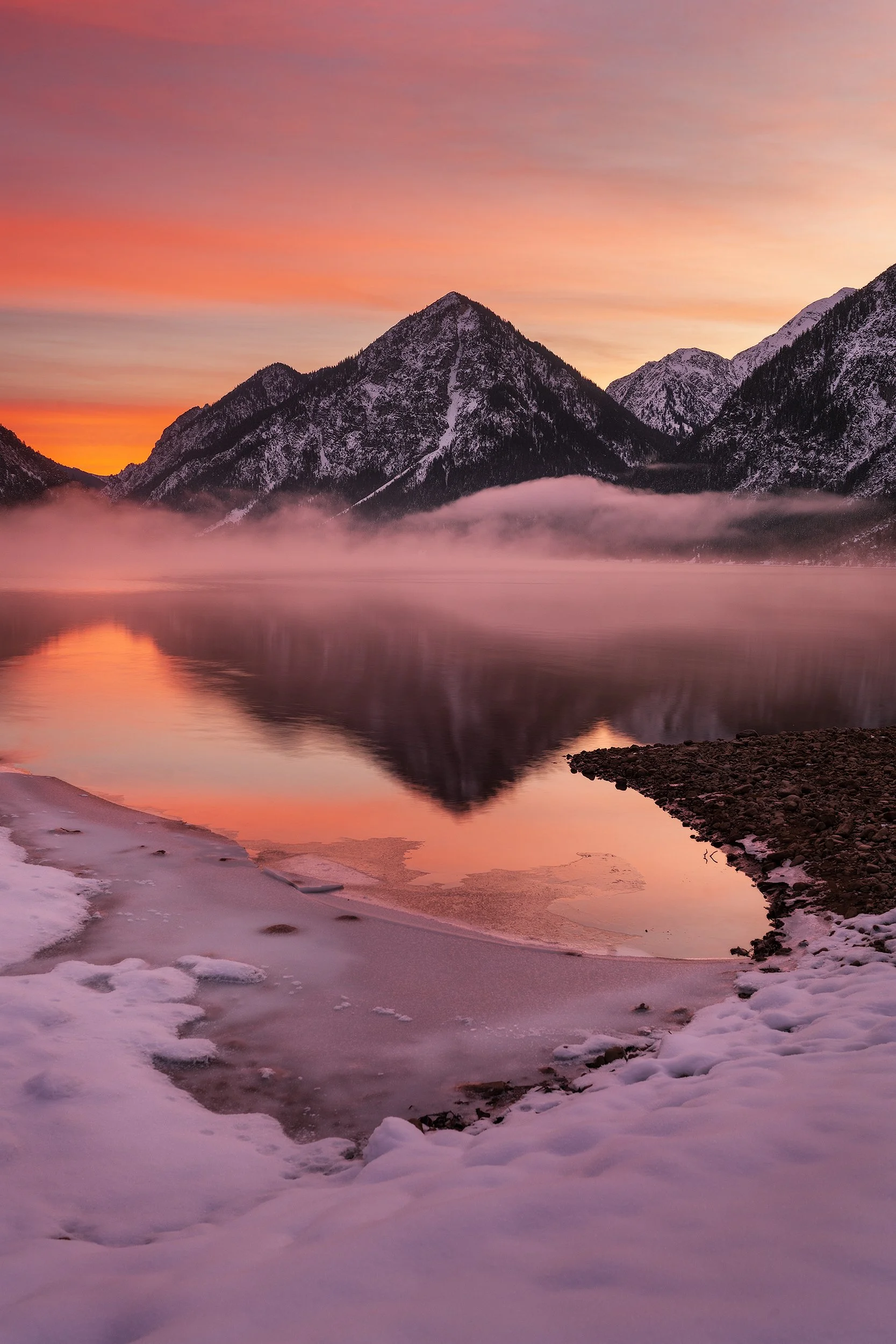 Serene mountain landscape with snow-capped peaks, calm lake reflecting the mountains, pink and orange sunset sky, and snow-covered shoreline.