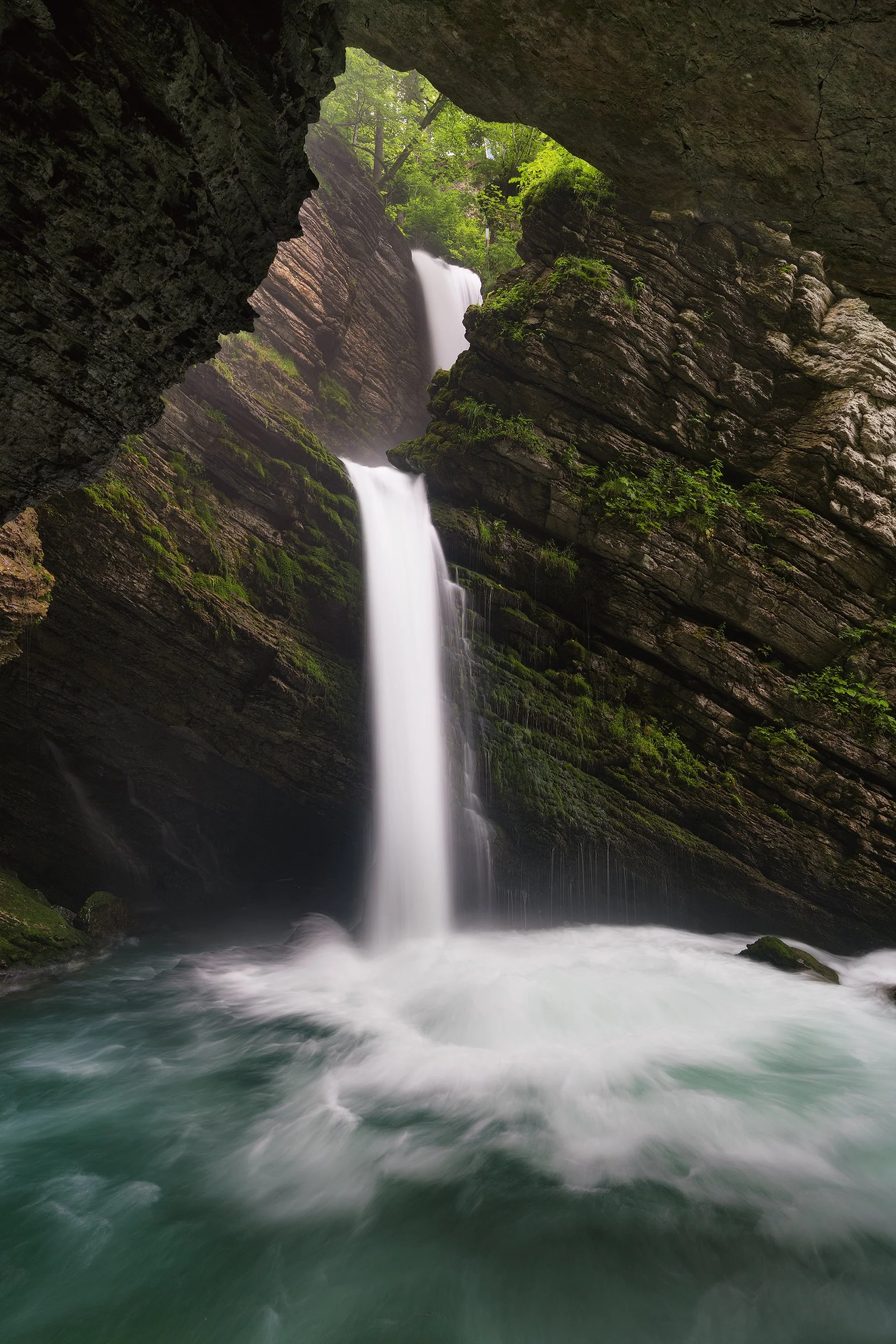 Waterfall cascading down rocks into a pool, viewed from inside a cave opening with trees above.