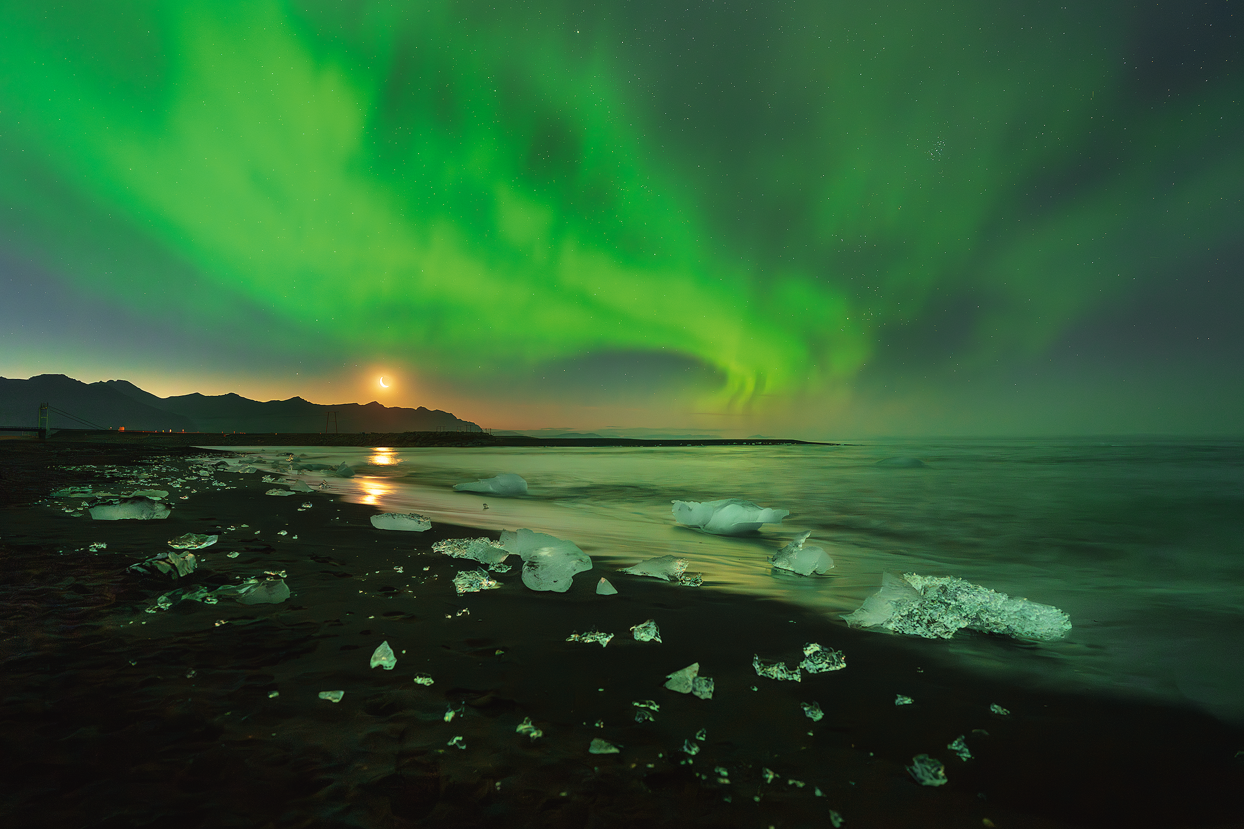 Northern lights over a dark beach with ice chunks, mountains in the background, and a crescent moon in the sky.