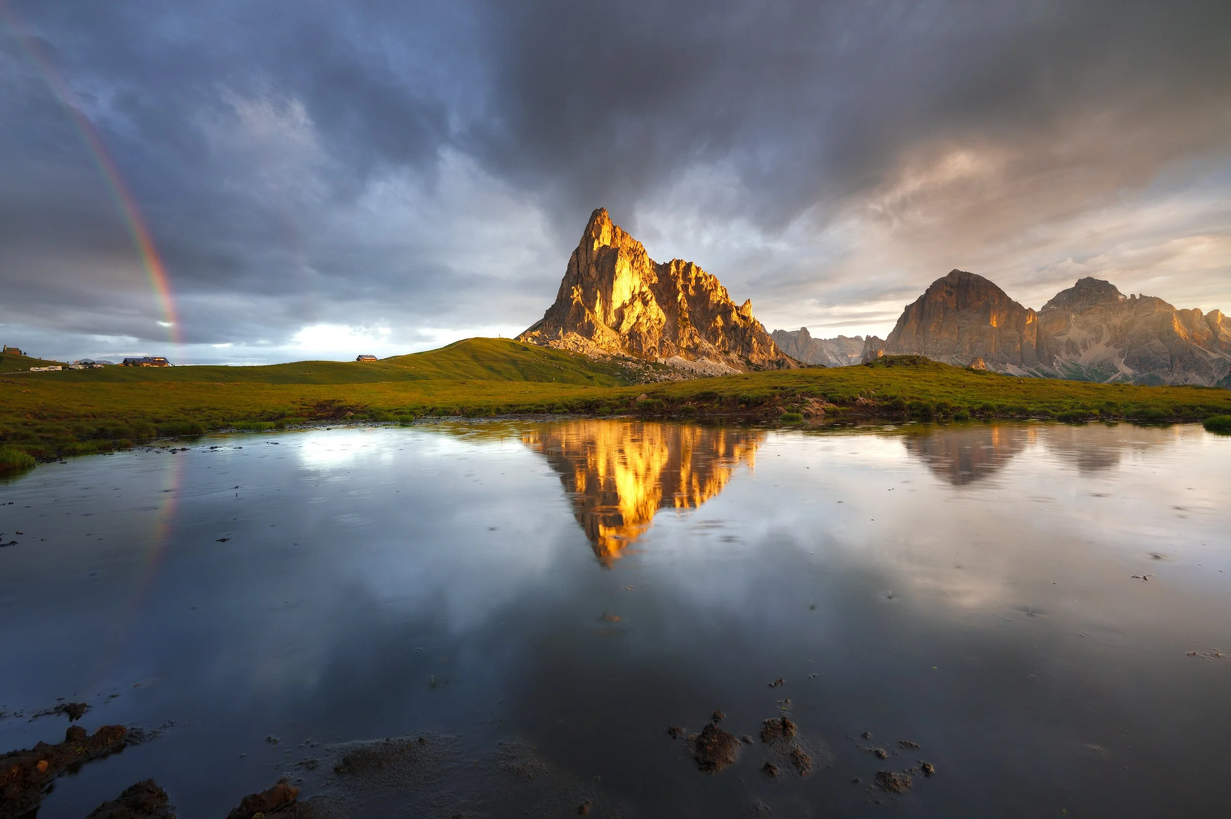 Mountain landscape with rocky peaks, green grass, a body of water reflecting the mountains, and a rainbow in a cloudy sky.