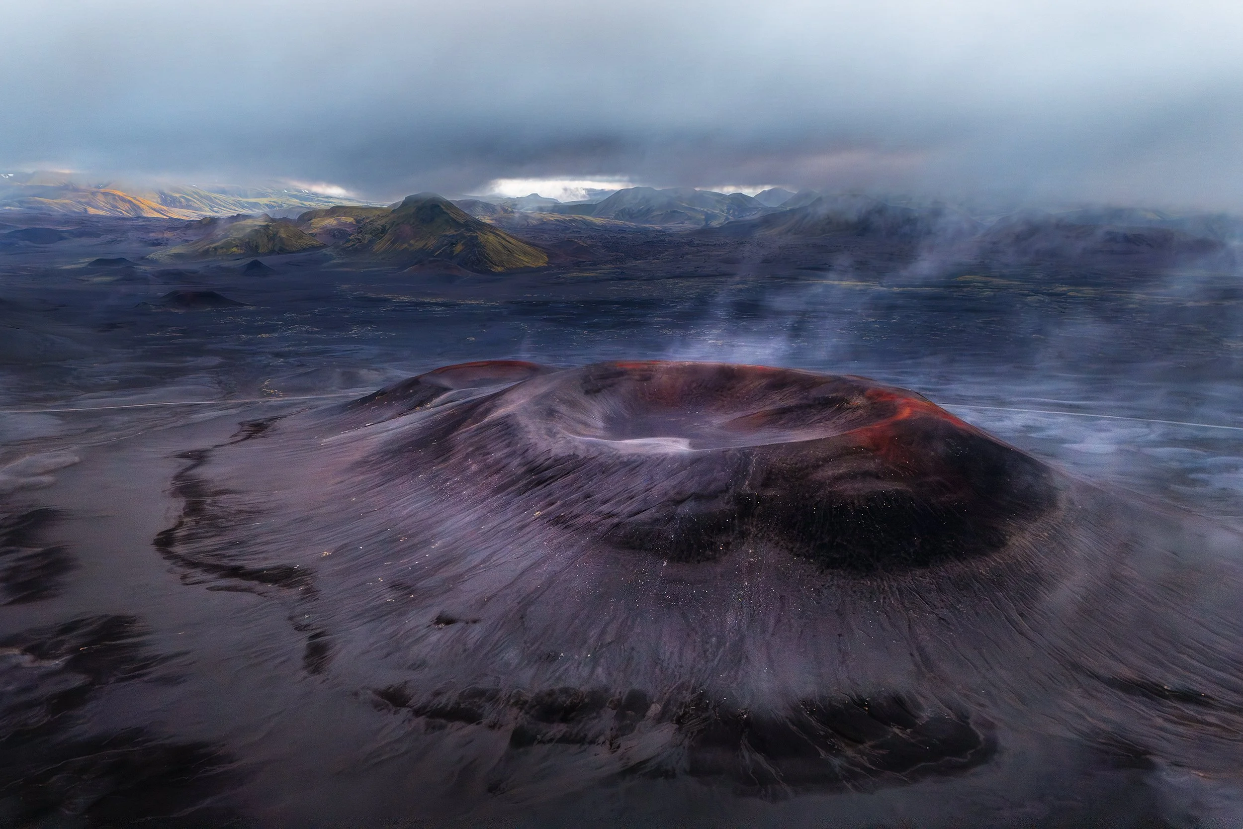 A volcanic landscape with a dormant volcano and smoke rising from its crater, surrounded by dark lava fields and distant mountains under a cloudy sky.