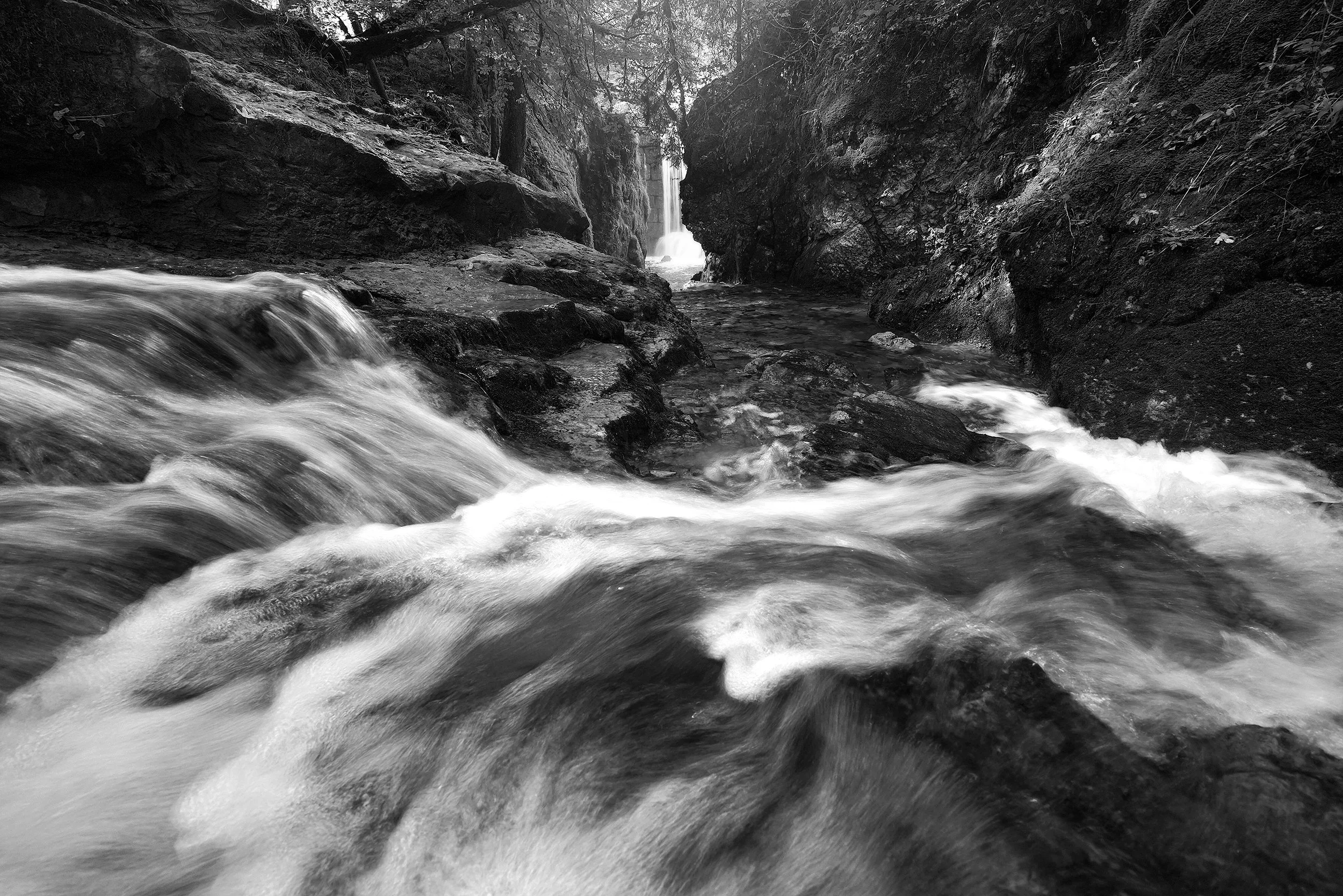 Black and white photo of a fast-moving stream flowing through rocky terrain with a waterfall in the background.