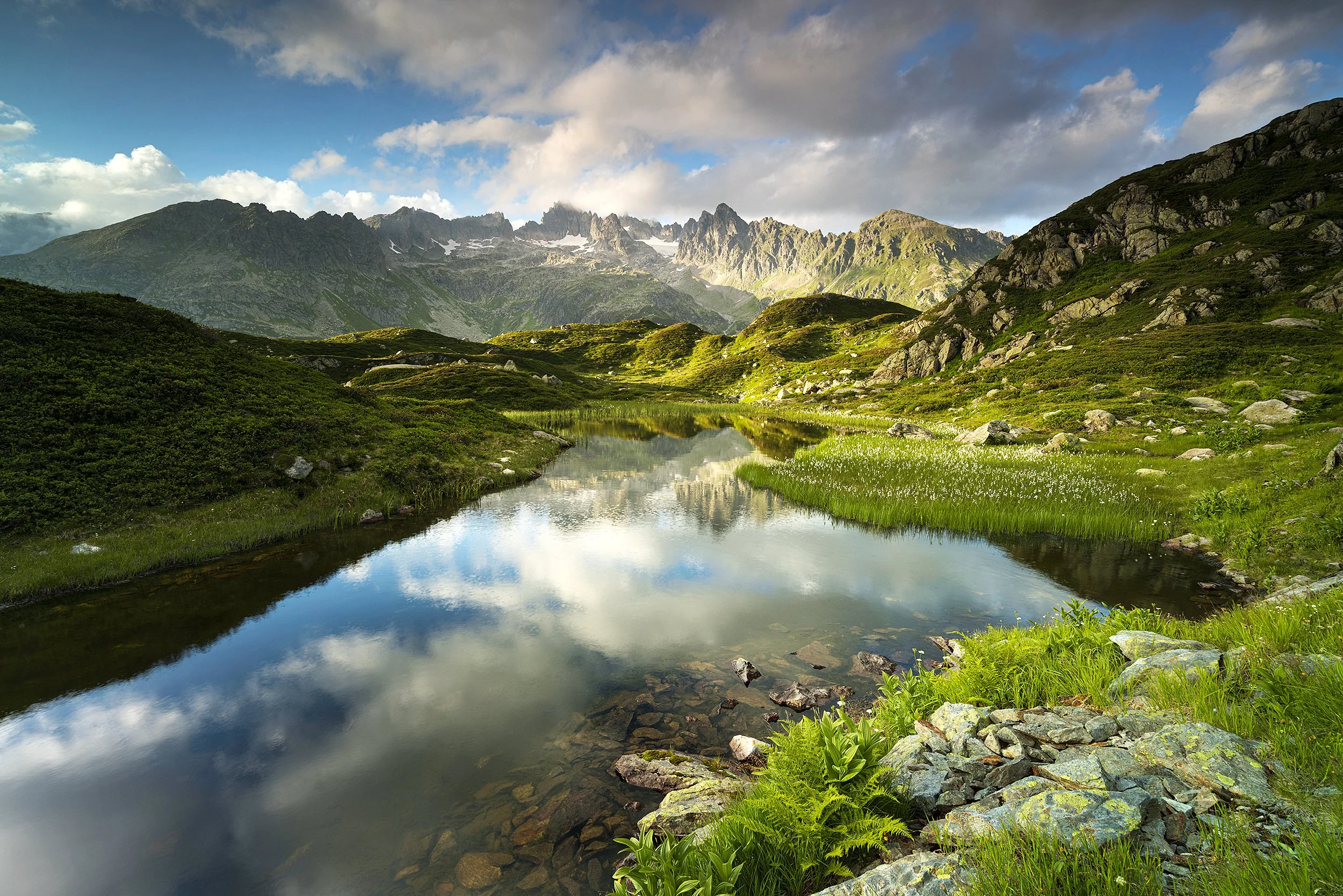 A scenic mountain landscape with a calm reflective river, lush green vegetation, rocky terrain, and towering mountains under a partly cloudy sky.