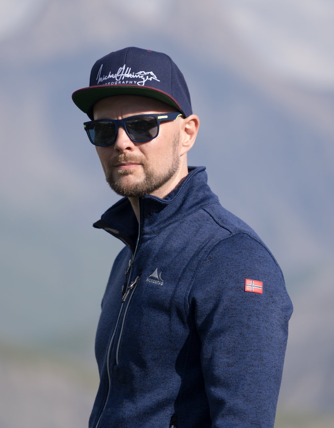 A man outdoors wearing a navy blue jacket, black sunglasses, a navy blue cap with white writing, and a Norwegian flag patch on his sleeve, against a cloudy sky background.