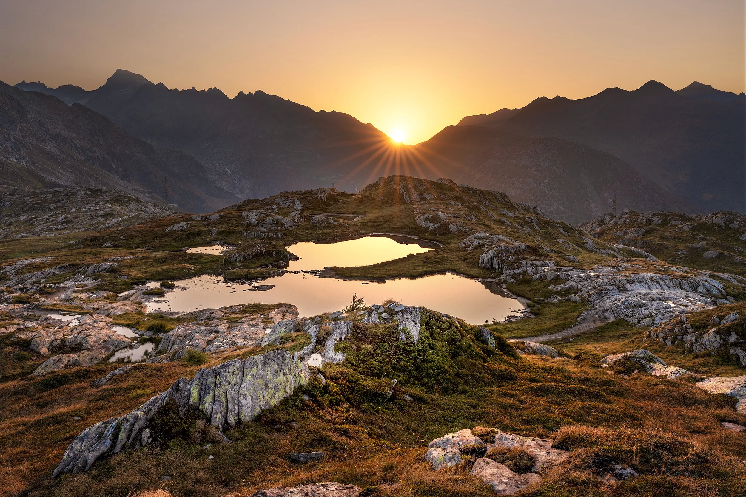 Sun setting behind mountain range illuminating the sky in warm hues, with rocky terrain and small water bodies in the foreground.