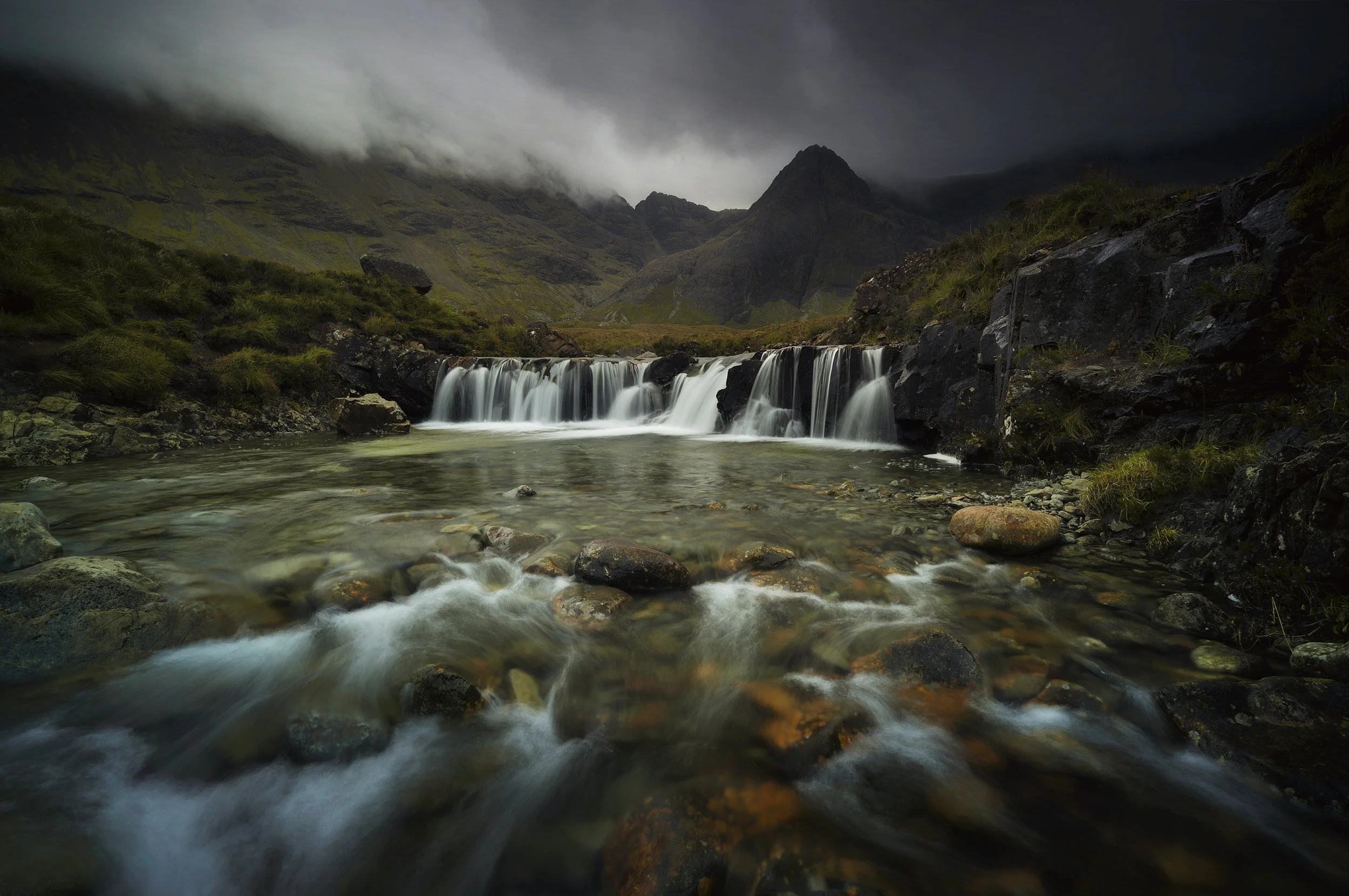 A mountain landscape with a small waterfall flowing into a rocky river under a cloudy sky.