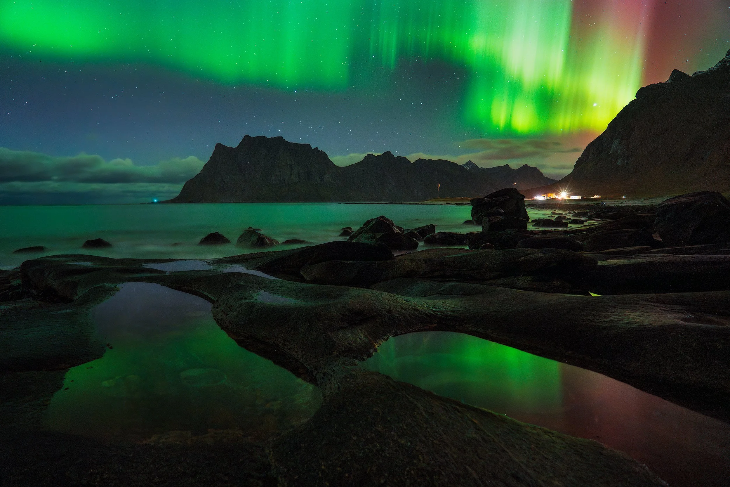 Northern Lights over a rocky shoreline at night with mountains in the background and a few lights from a building.