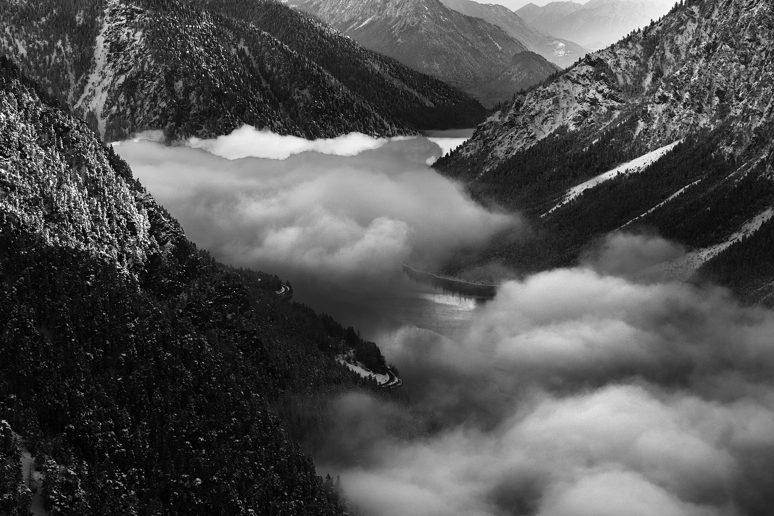 A black and white photograph of a mountain landscape with steep, rugged, snow-capped mountains and a winding river or lake below, surrounded by mist or fog.