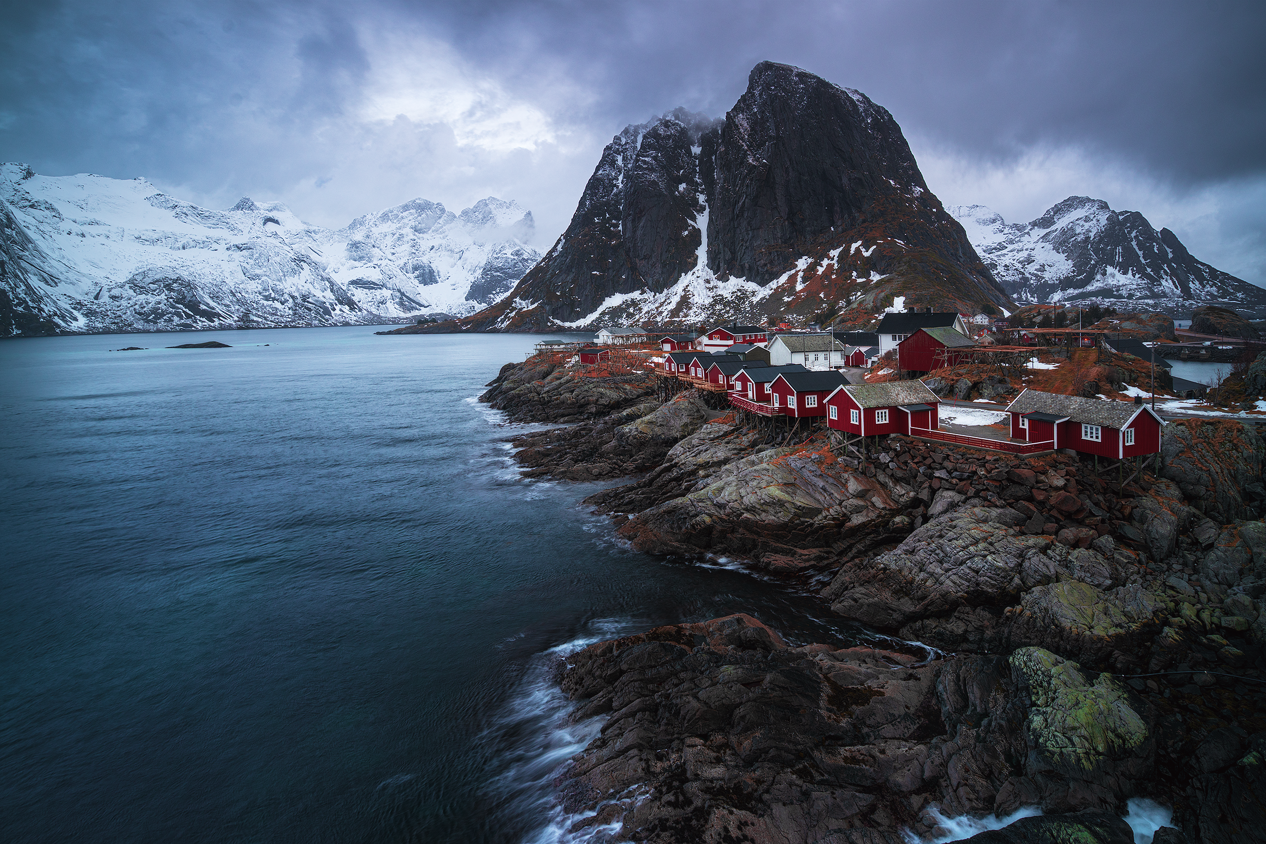 Coastal village with red houses on rocky shoreline, large mountain with snow in background, overcast sky.