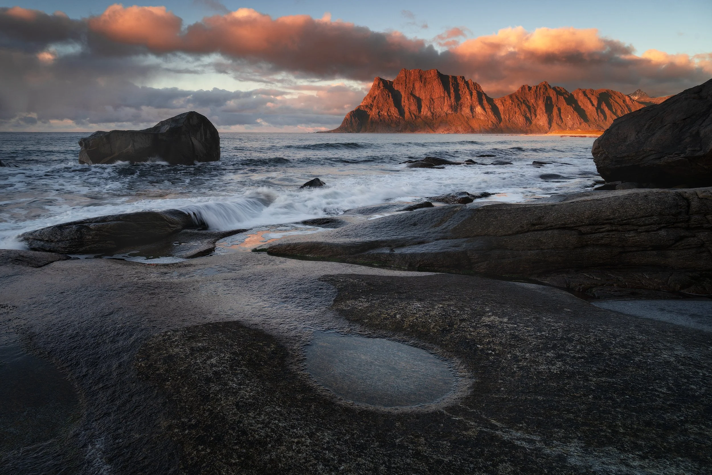 A rocky seashore at sunset with waves crashing against rocks, a large distant mountain illuminated in orange, and a sky with clouds.