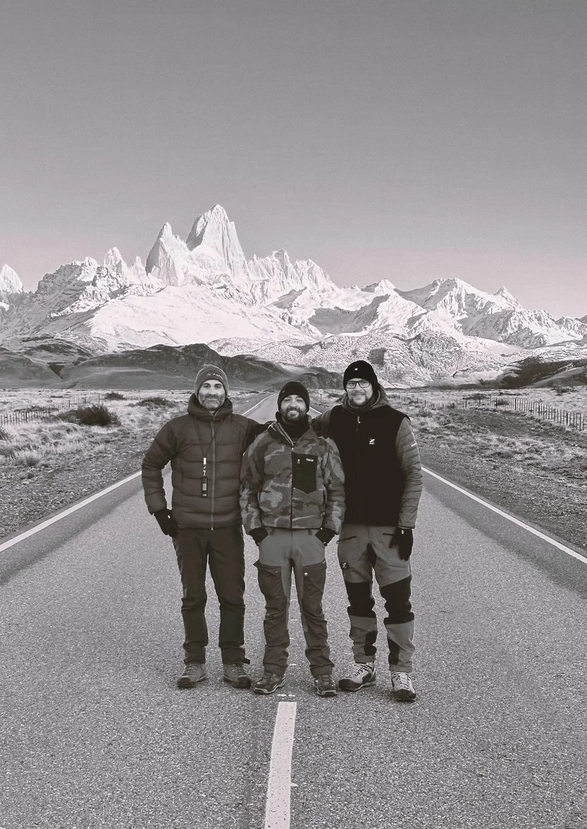 Three men standing on a road with snow-covered mountains in the background.