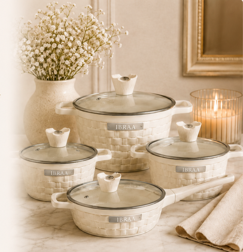Set of four ivory-colored ceramic cookware with glass lids and ivory knobs, labeled 'IBRAA,' placed on a marble surface with a vase of white flowers and a lit candle in the background.