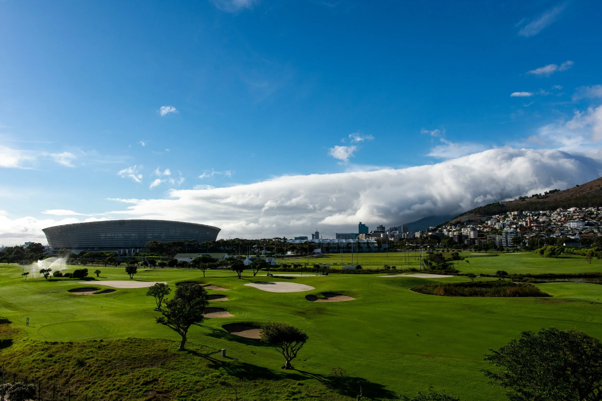 A golf course with sand traps, green grass, trees, and a fountain, with a large stadium and cityscape in the background under a partly cloudy sky.