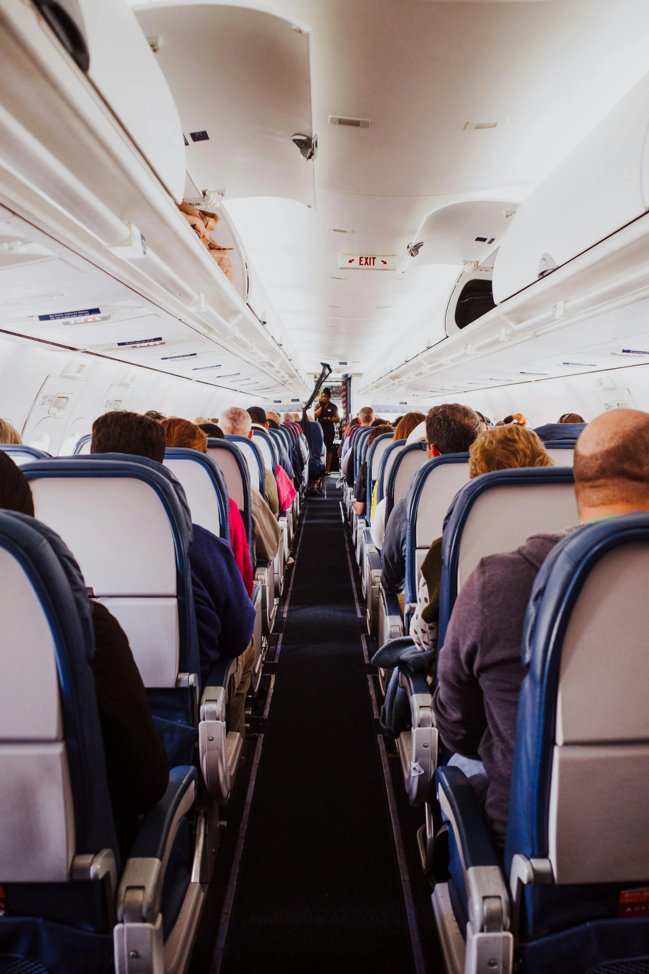 View inside an airplane cabin showing rows of passengers seated, with overhead compartments above, and an instructor at the front of the plane, possibly conducting a safety demonstration.