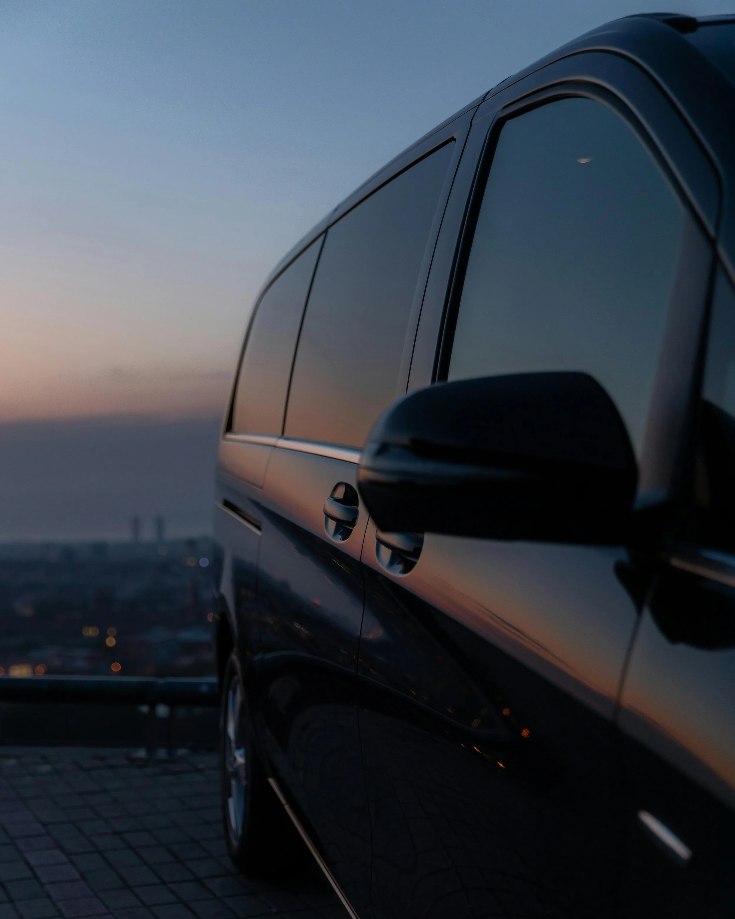 Close-up of a black vehicle with tinted windows parked on a tiled surface at dusk, with a city skyline blurred in the background.