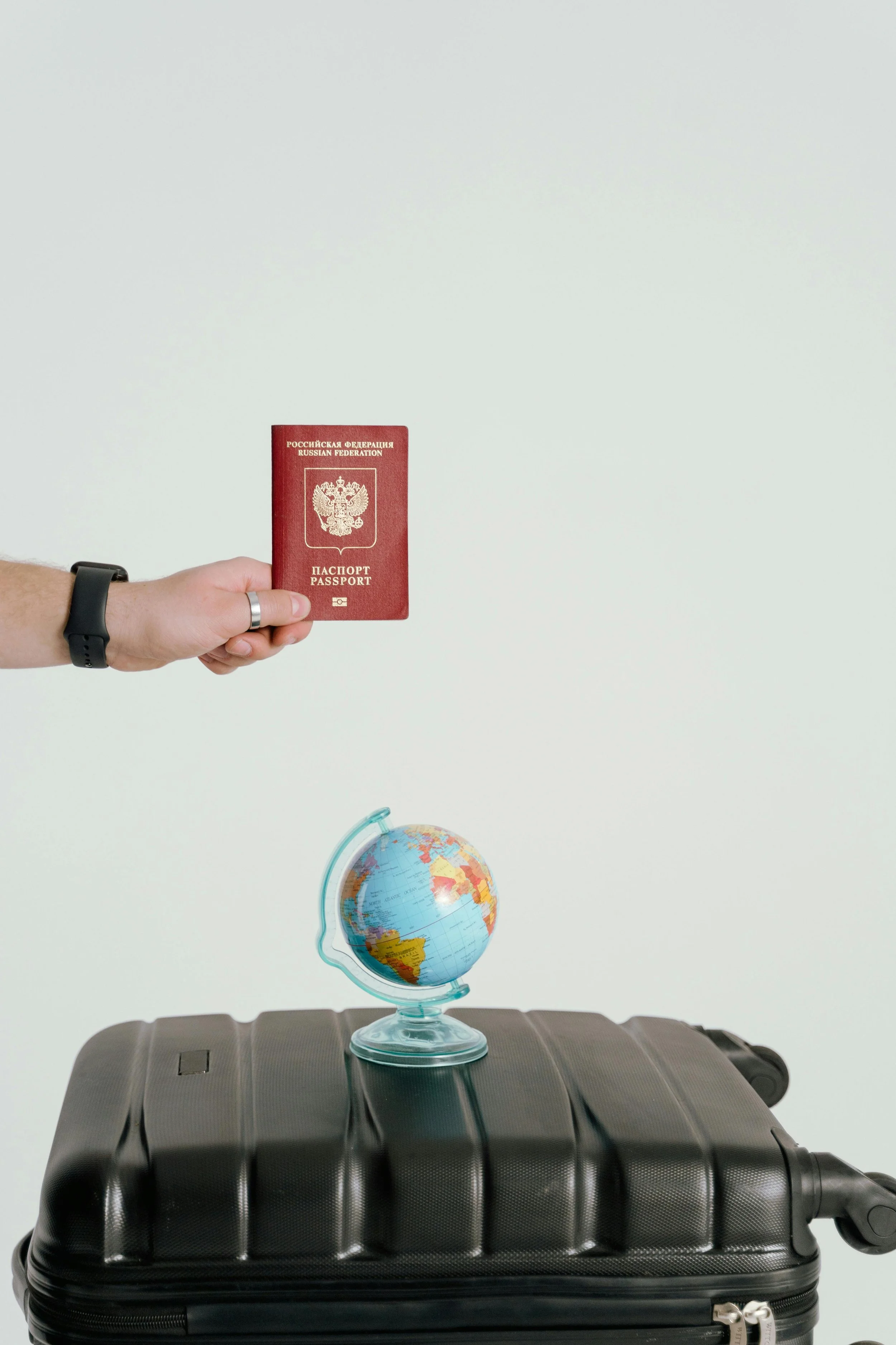 A hand with a wristwatch holding a Russian Federation passport above a black suitcase, with a globe on a table below.