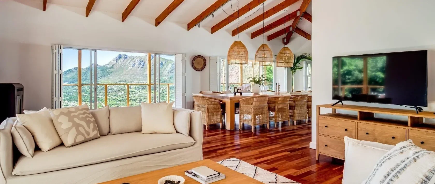 Living room with white sofa, wooden coffee table, and a flat-screen TV on a wooden stand. Behind is a dining area with a large wooden table and wicker chairs, pendant lights overhead, and large windows showing mountain views.