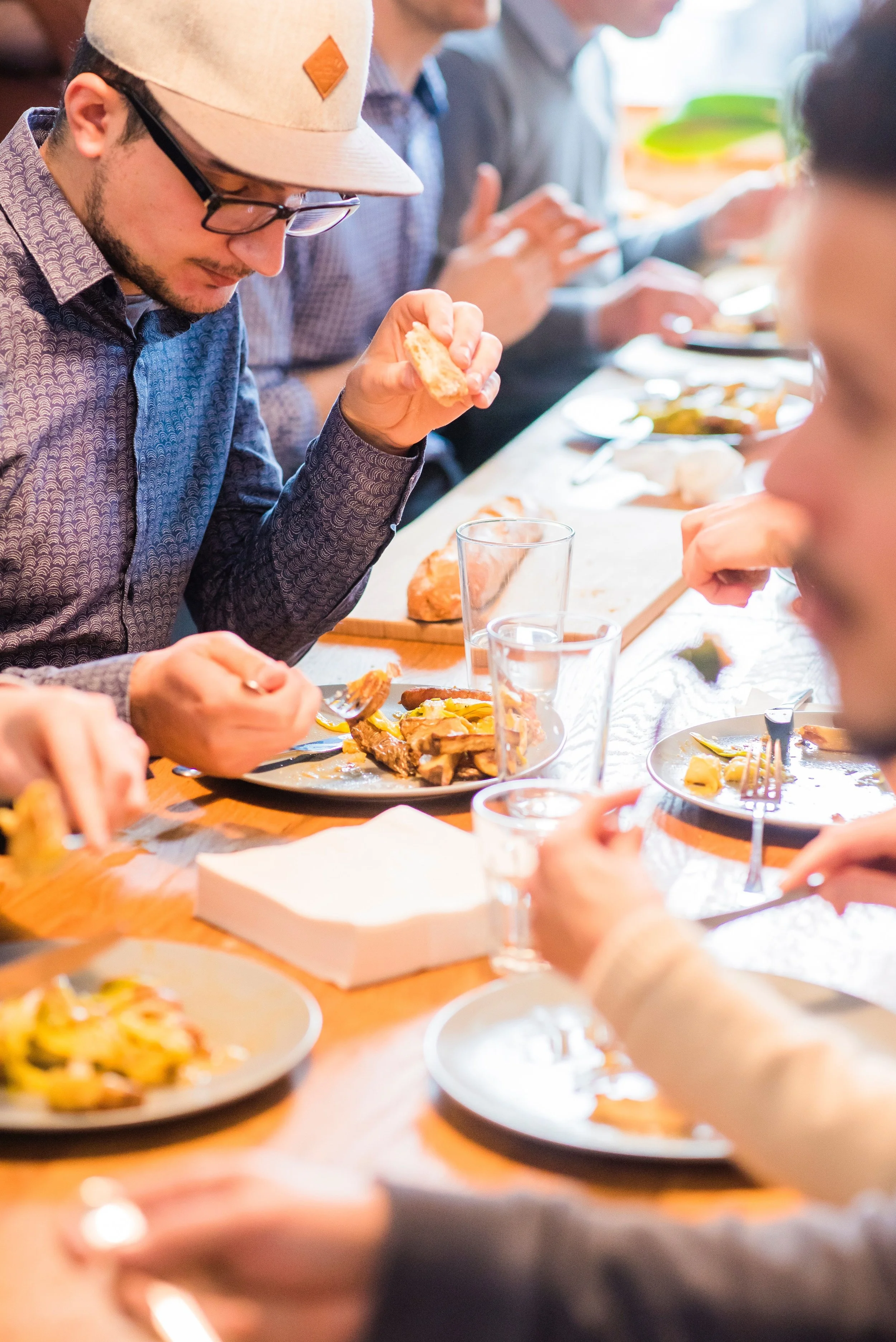 A group of people sitting at a wooden table enjoying a meal together, with plates of food, glasses, and utensils.