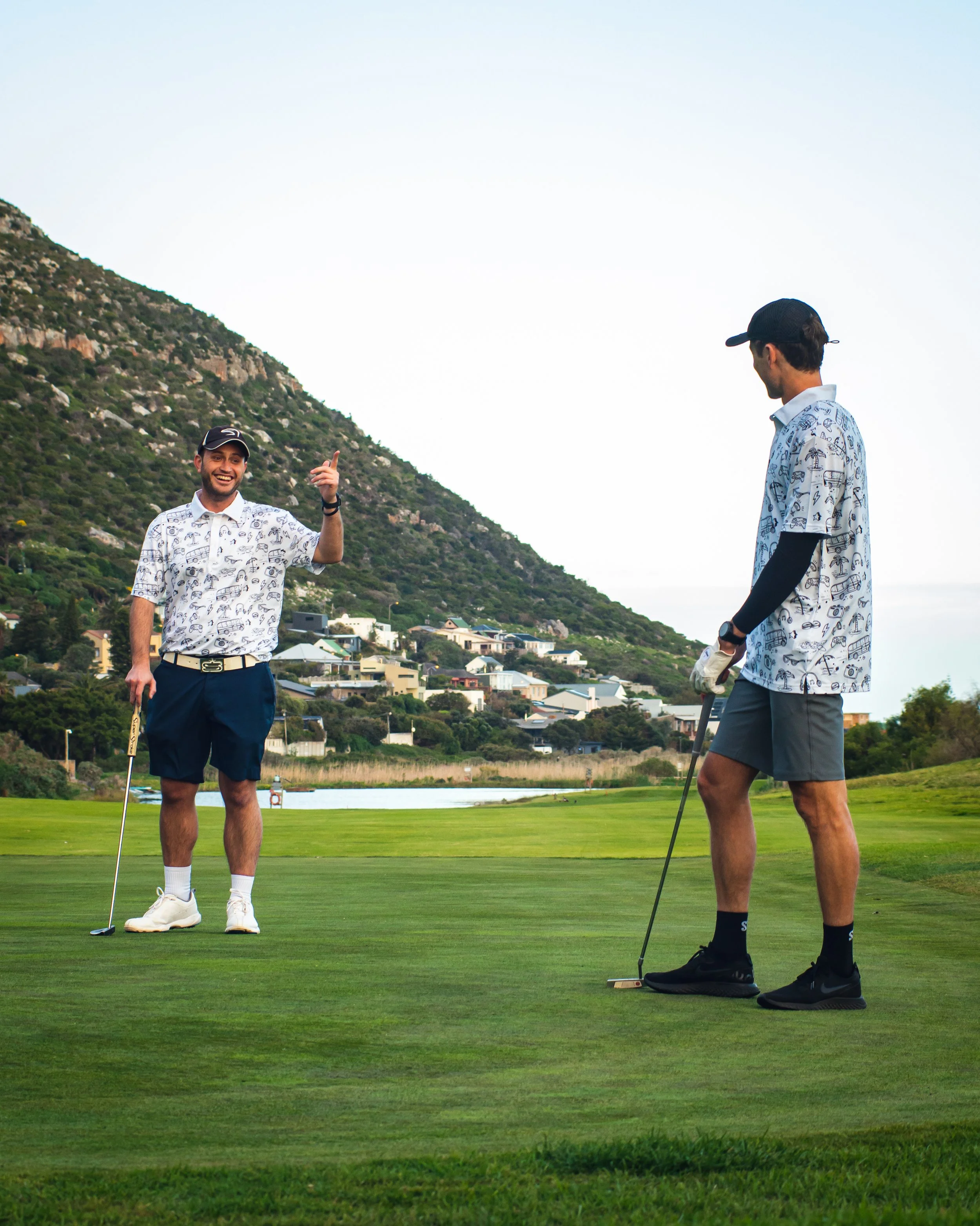 Two men playing golf on a course with hills and houses in the background, one smiling and pointing, the other preparing to putt.