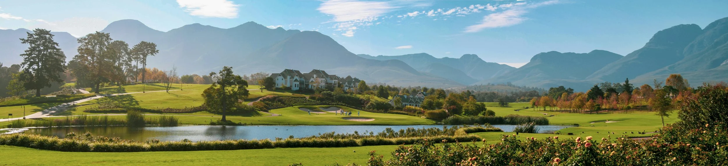 Scenic view of a golf course with a pond, trees, pathways, residential buildings, and mountain range in the background on a clear day.
