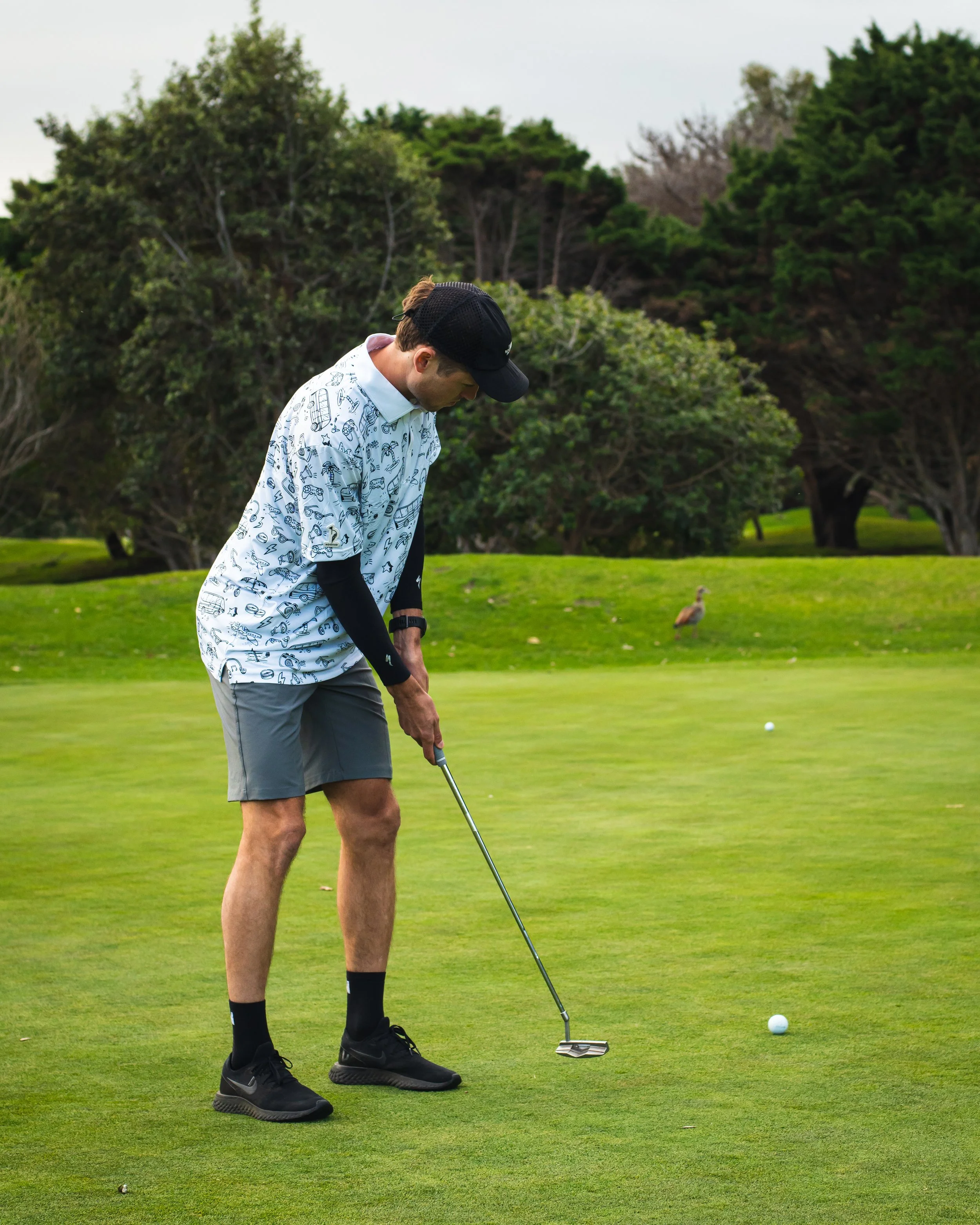 Man in a patterned white shirt, gray shorts, and black cap preparing to putt on a golf course. He is holding a golf club, and a golf ball is on the green in front of him. There are trees and a duck in the background.